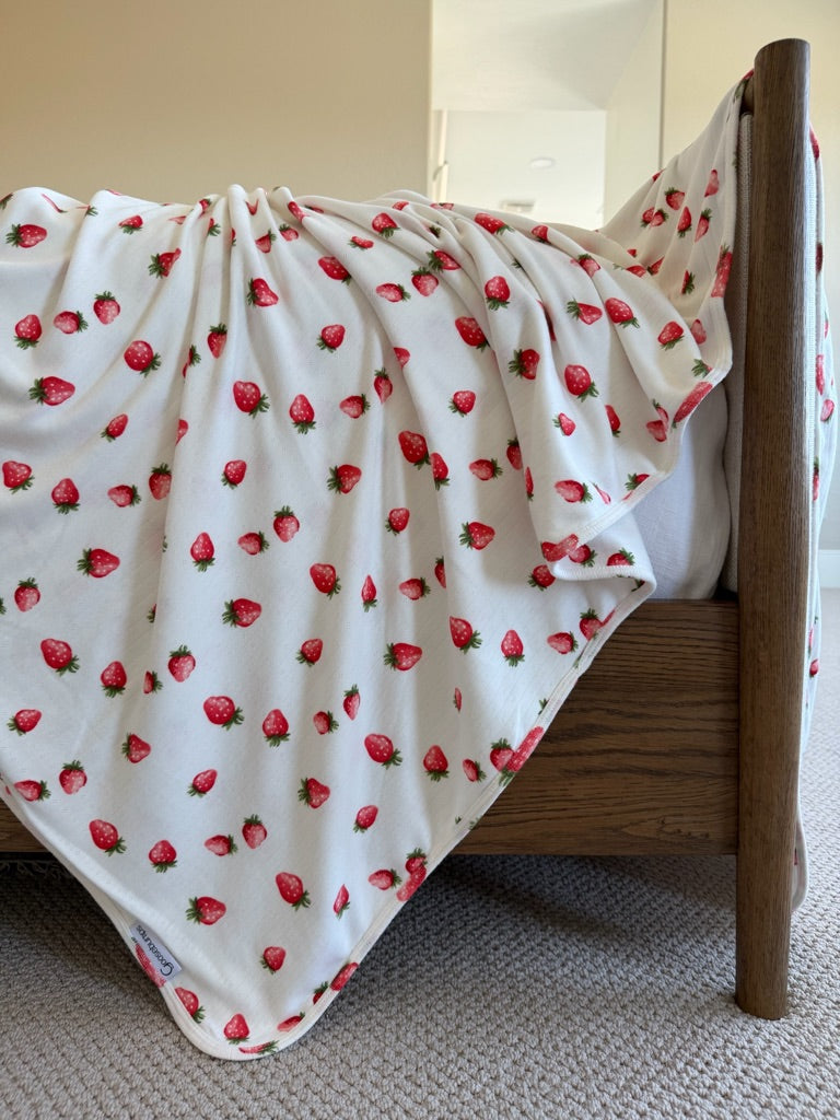 The berries / pointelle blanket with a red strawberry print is draped over the edge of a wooden bed in a bright, carpeted bedroom.