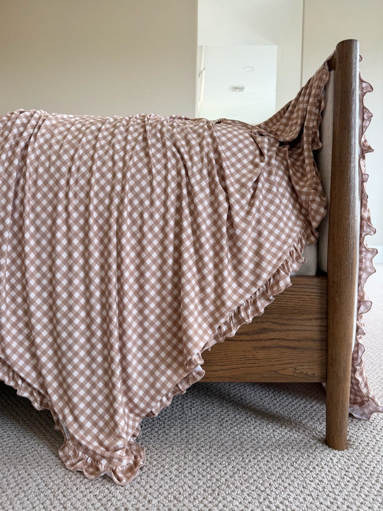 The teddy bear check / ruffle blanket in brown and white gently drapes over the side of a wooden bed frame, its soft ruffles cascading onto a beige carpeted floor in a softly lit room.