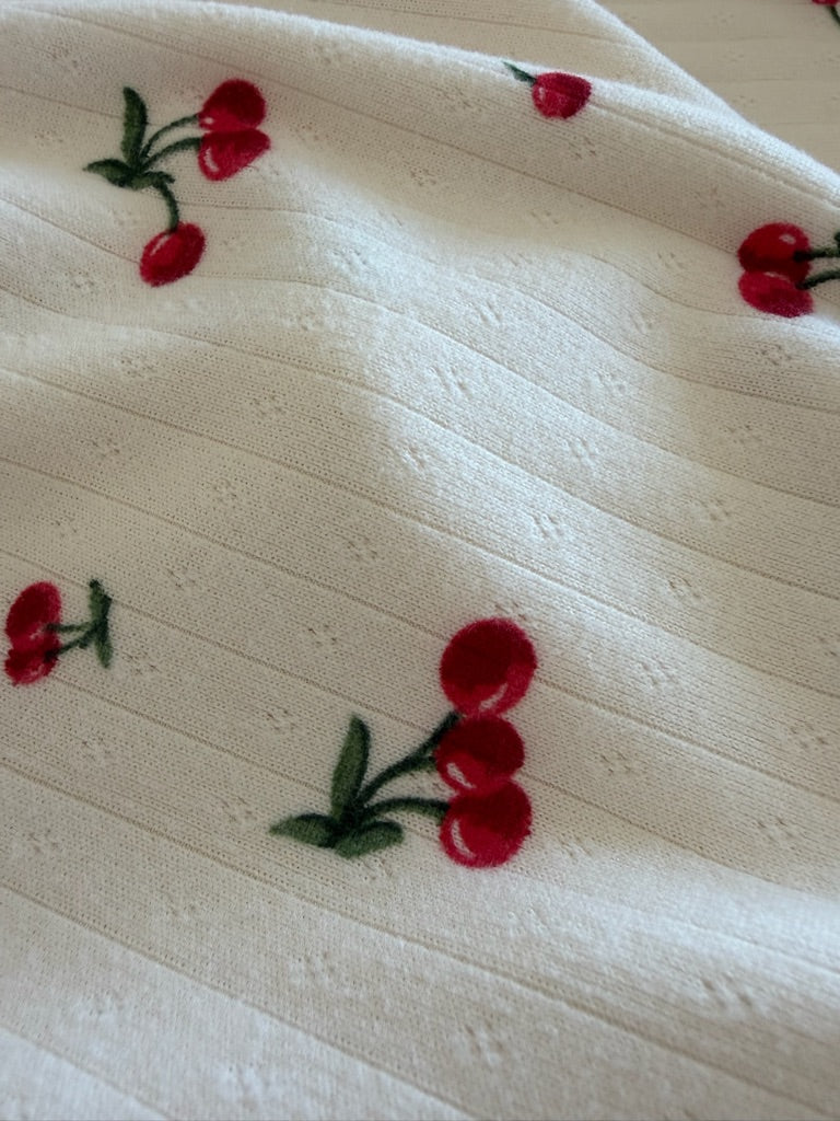 A close-up of shirley temple / pointelle shows white, ribbed fabric adorned with small red cherries and green stems, giving a playful cherry print look with a soft texture.