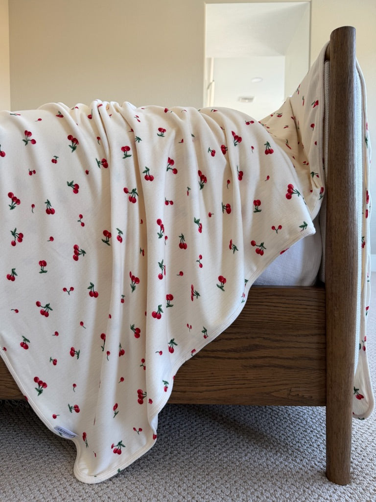 The shirley temple / pointelle blanket in cream with a cherry print is draped over the side of a wooden bed in a softly lit room with beige walls and a carpeted floor.