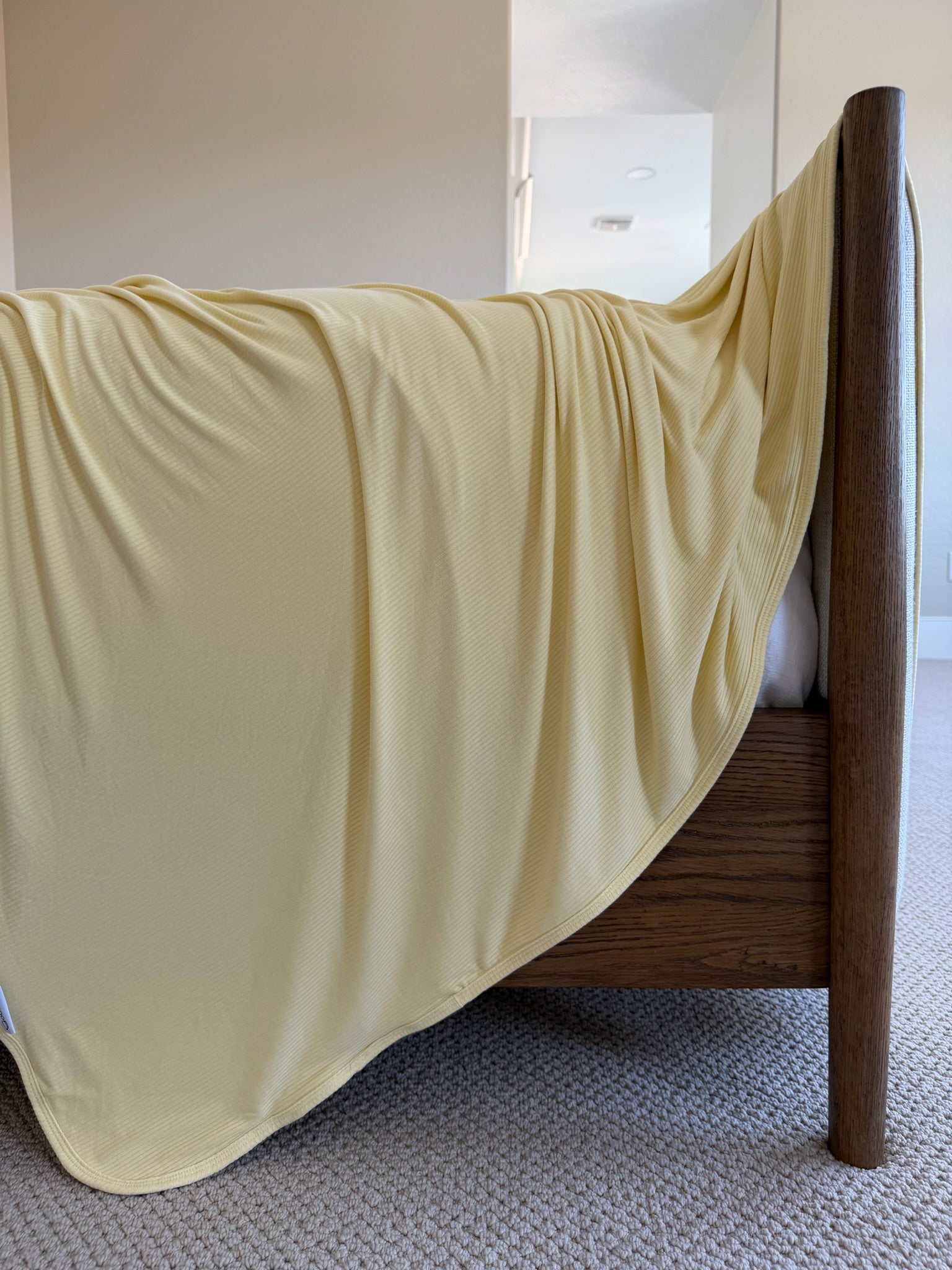 A buttery-soft bamboo blanket in light yellow called "butter" drapes over the wooden footboard of a bed in a cozy, carpeted room filled with gentle natural light and neutral-colored walls.