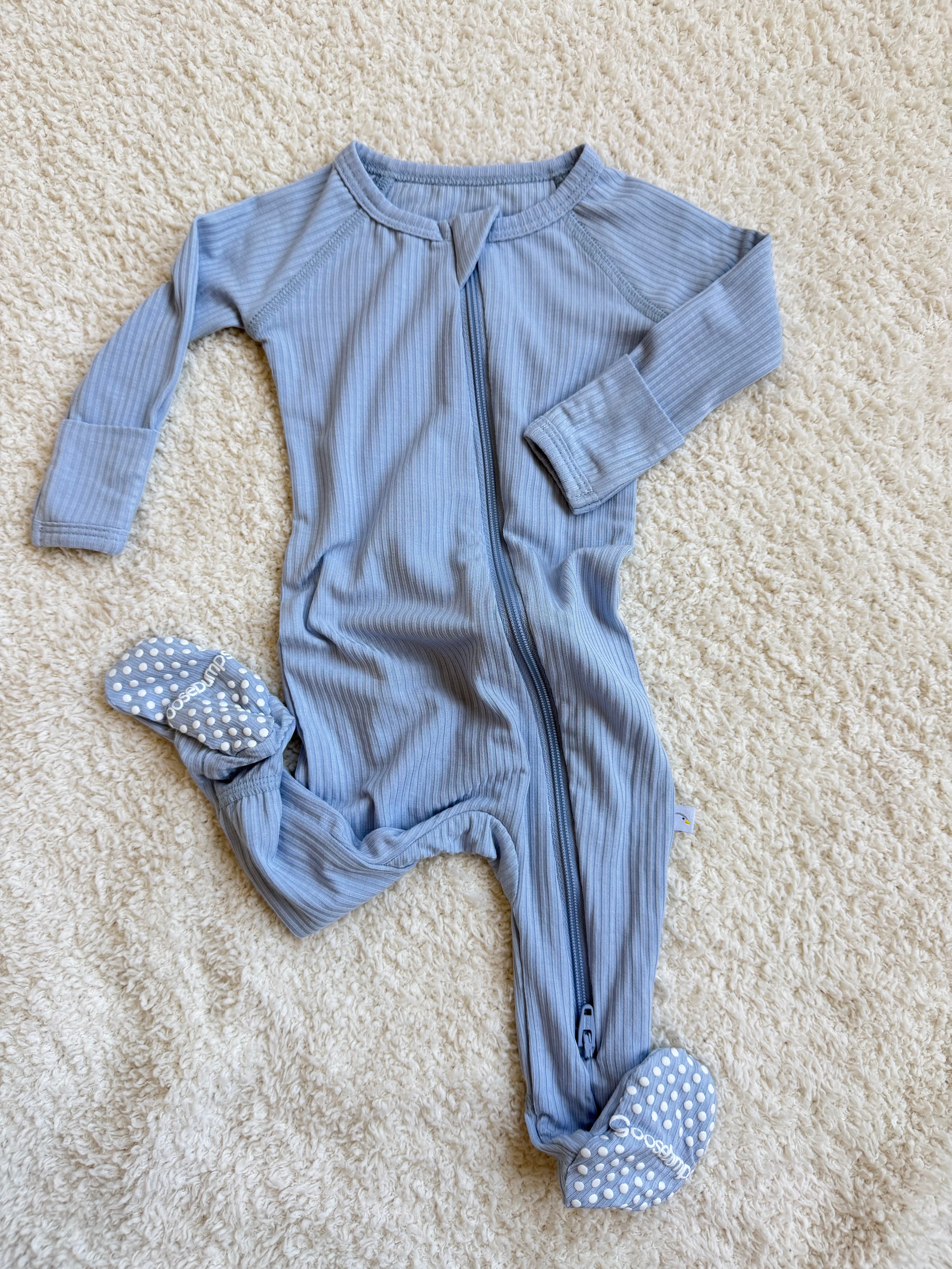 A cove one piece zipper, featuring long sleeves and a light blue ribbed fabric with attached polka-dot footies, lies on a cream-colored textured surface.