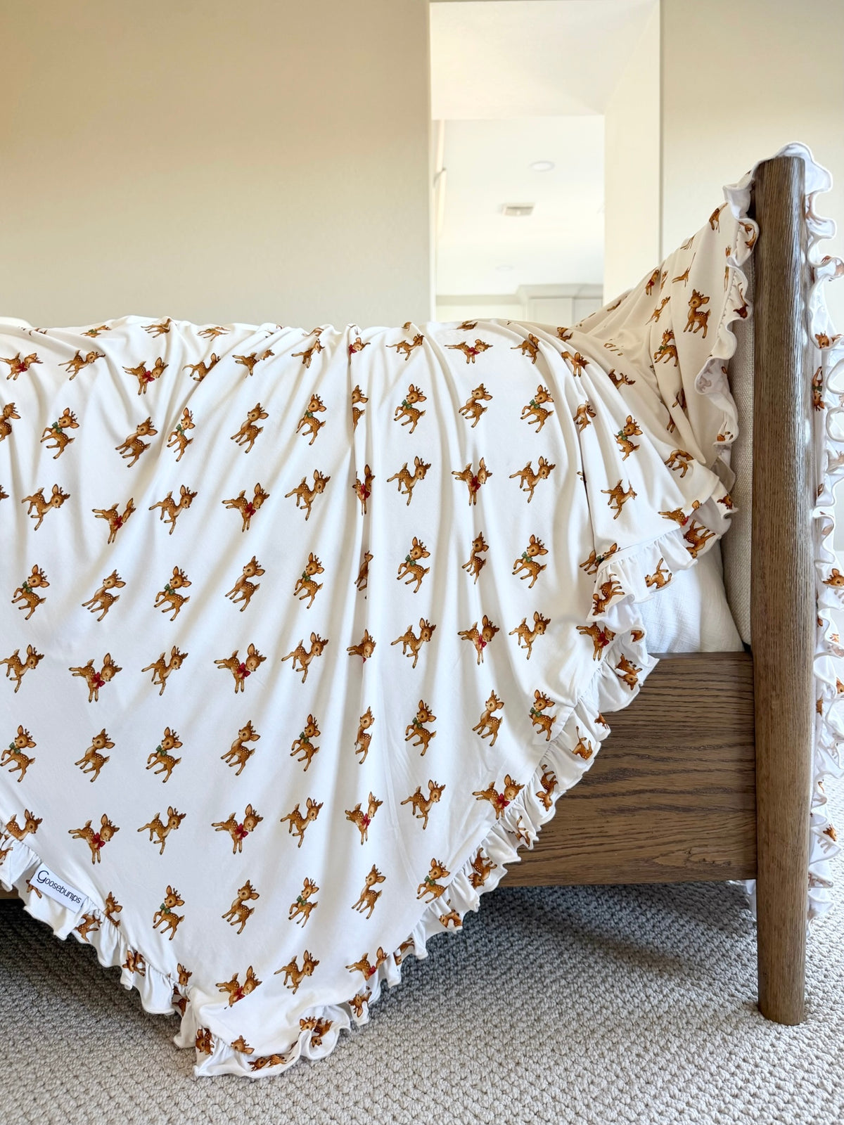 Vintage reindeer / ruffle baby blanket in white with a ruffled edge and repeating reindeer print, draped over the side of a wooden bed.