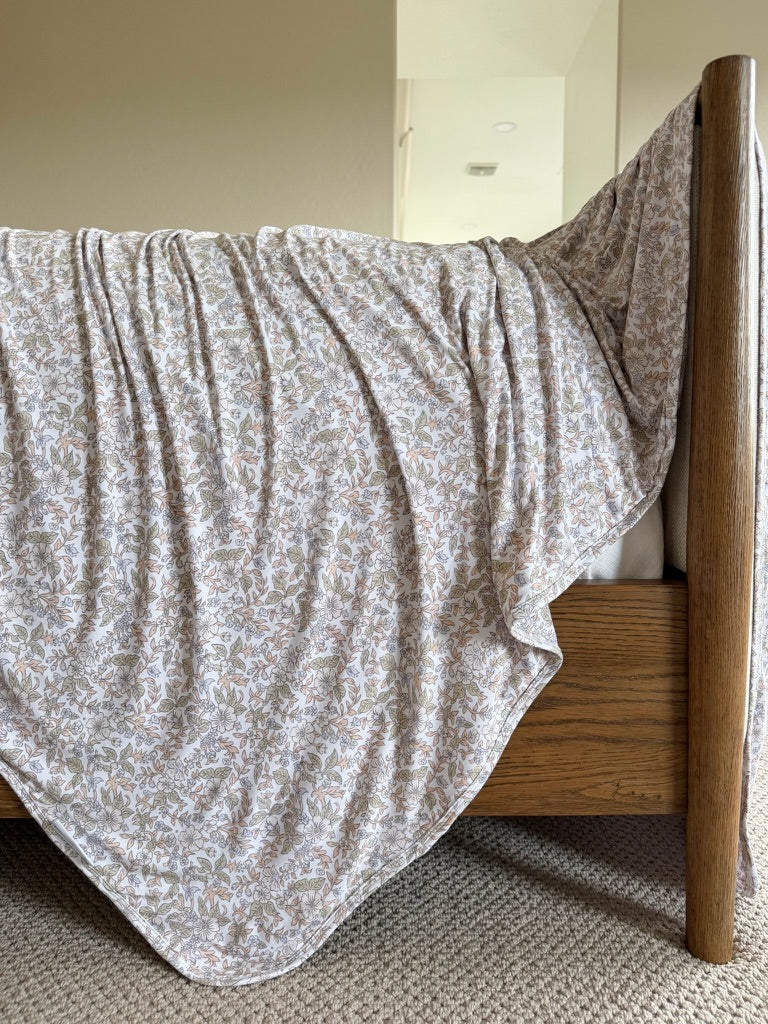 An heirloom blanket with a floral print is draped over the wooden footboard of a bed in a carpeted room with neutral-colored walls.