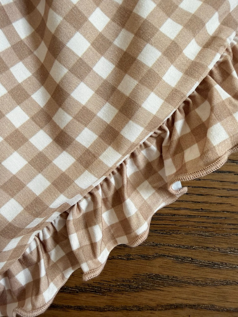 A close-up of the teddy bear check / ruffle blanket, featuring a beige and white gingham pattern with ruffled edges, resting on a wooden surface with visible grain patterns.
