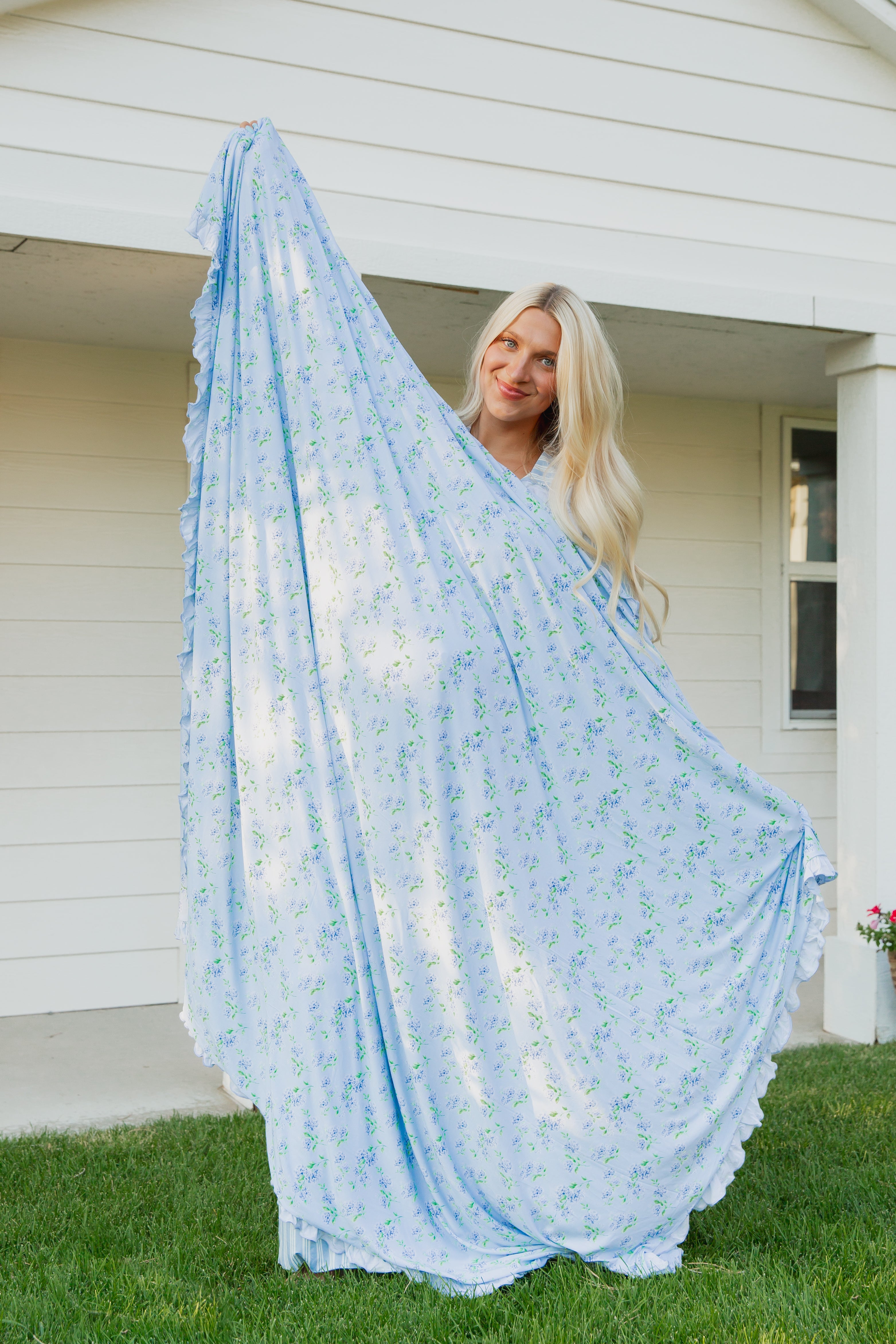 A woman with long blonde hair stands on grass, smiling and holding up the hydrangea / ruffle blanket—an oversized, luxurious bamboo throw featuring a light blue hydrangea print—in front of a white house.