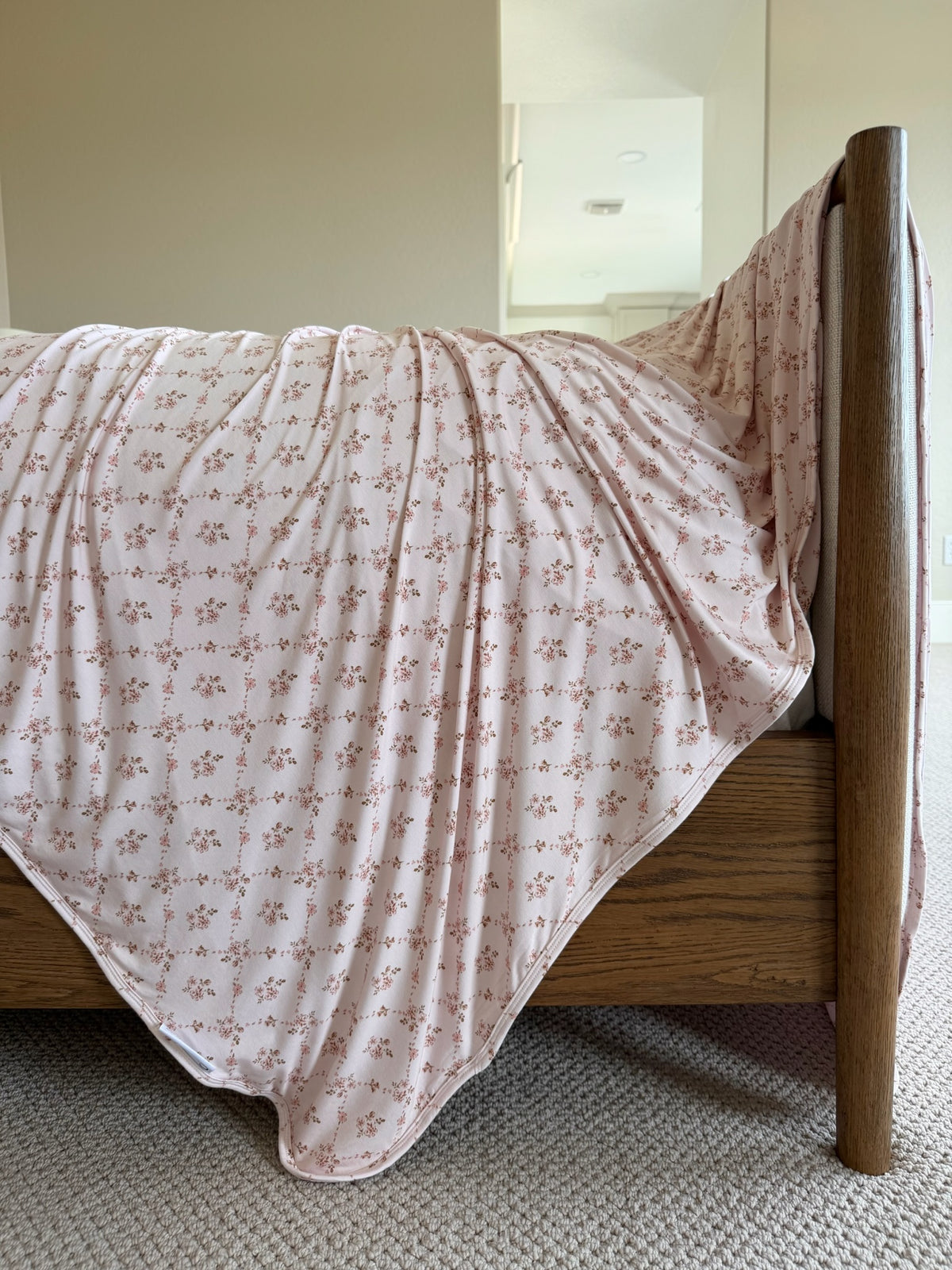A close-up of the keepsake wooden bed showcases a pink floral print blanket draped over the side, covering most of the bed. The room features beige carpeting and neutral-colored walls.