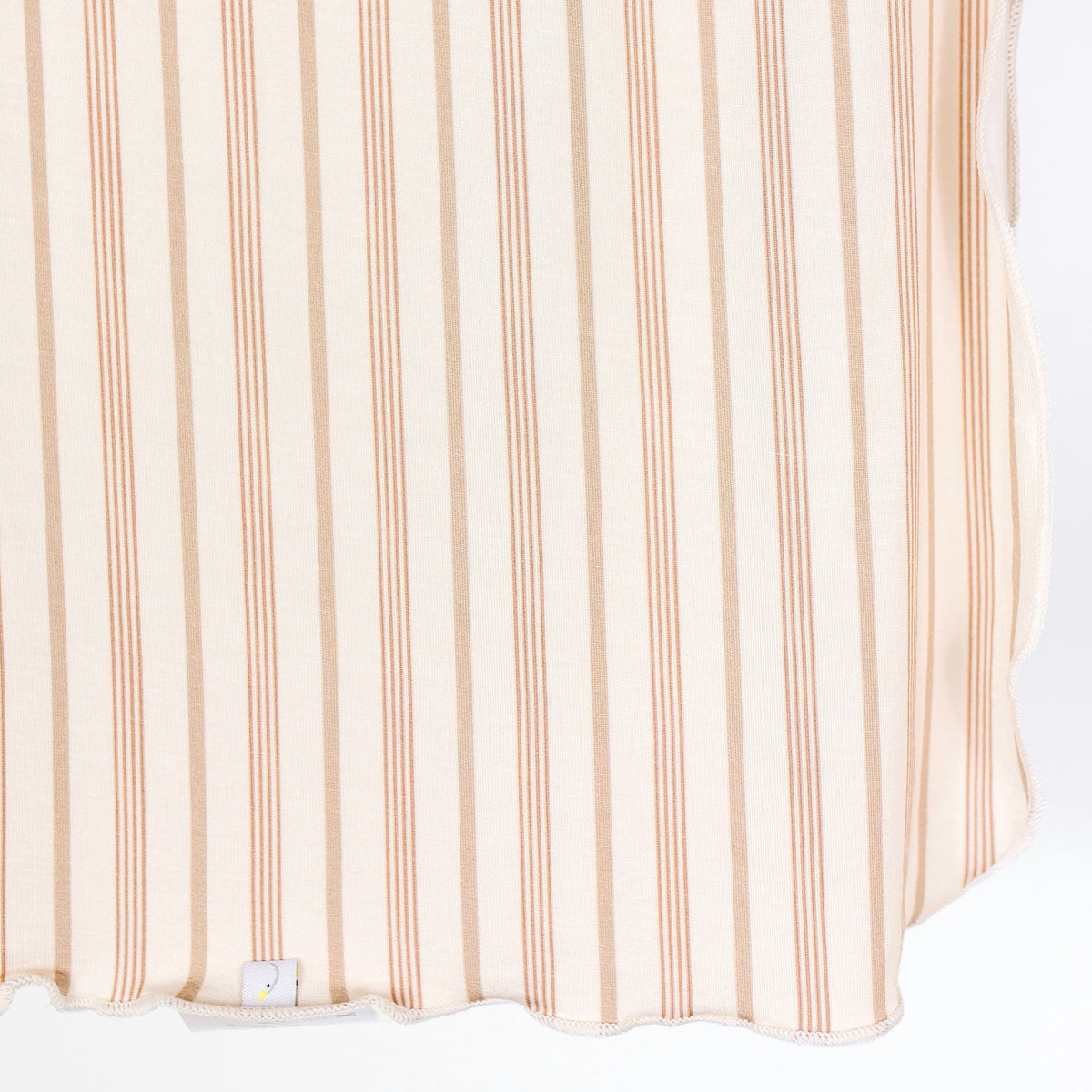 A close-up of beige fabric with vertical light brown and peach stripes, featuring a wavy scalloped edge and a small white tag near the bottom—this toast and honey / swaddle is crafted as a lightweight, single-layer baby blanket perfect for everyday comfort.