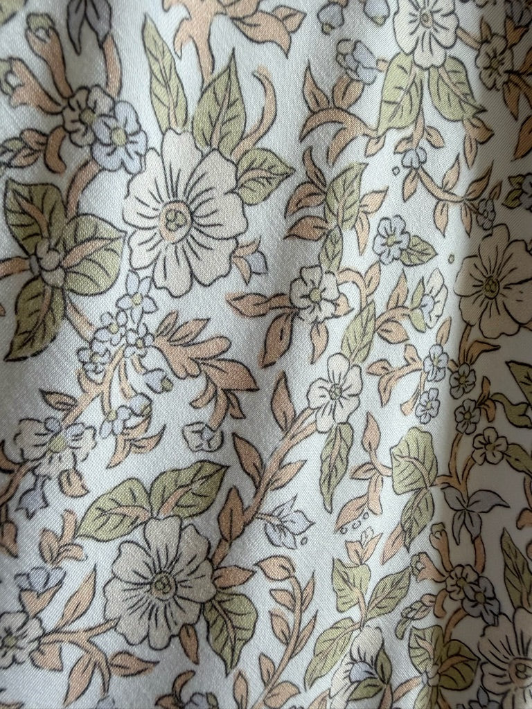 Close-up of the Heirloom blanket, featuring an elegant floral print with white and beige flowers, green leaves, and flowing vines set against a light background. The gently wrinkled fabric accentuates the soft texture and luxurious feel of this beautiful blanket.