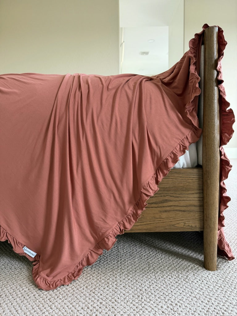 A close-up of a wooden bed adorned with the “serenity / ruffle” luxurious rust-colored ruffled blanket, draped elegantly over the side and resting on a beige carpeted floor in a softly lit room.
