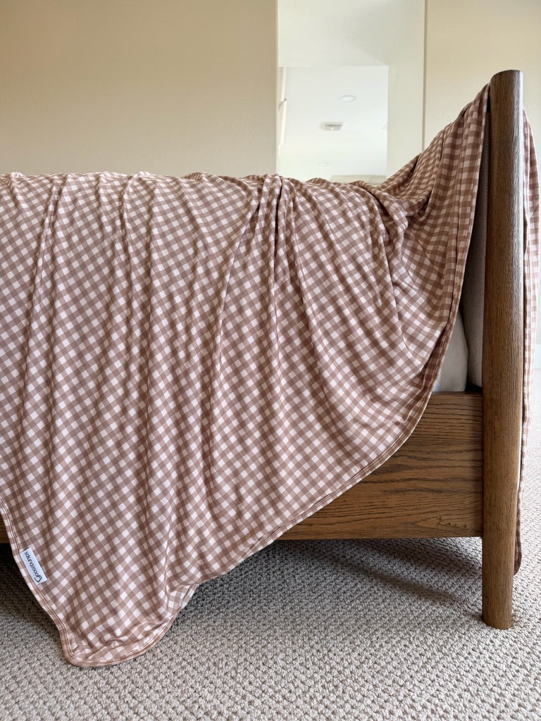 The “teddy bear check” blanket, featuring a brown and white gingham pattern, is draped over the side of a wooden bed frame, complemented by beige carpeting and neutral-colored walls in the background.