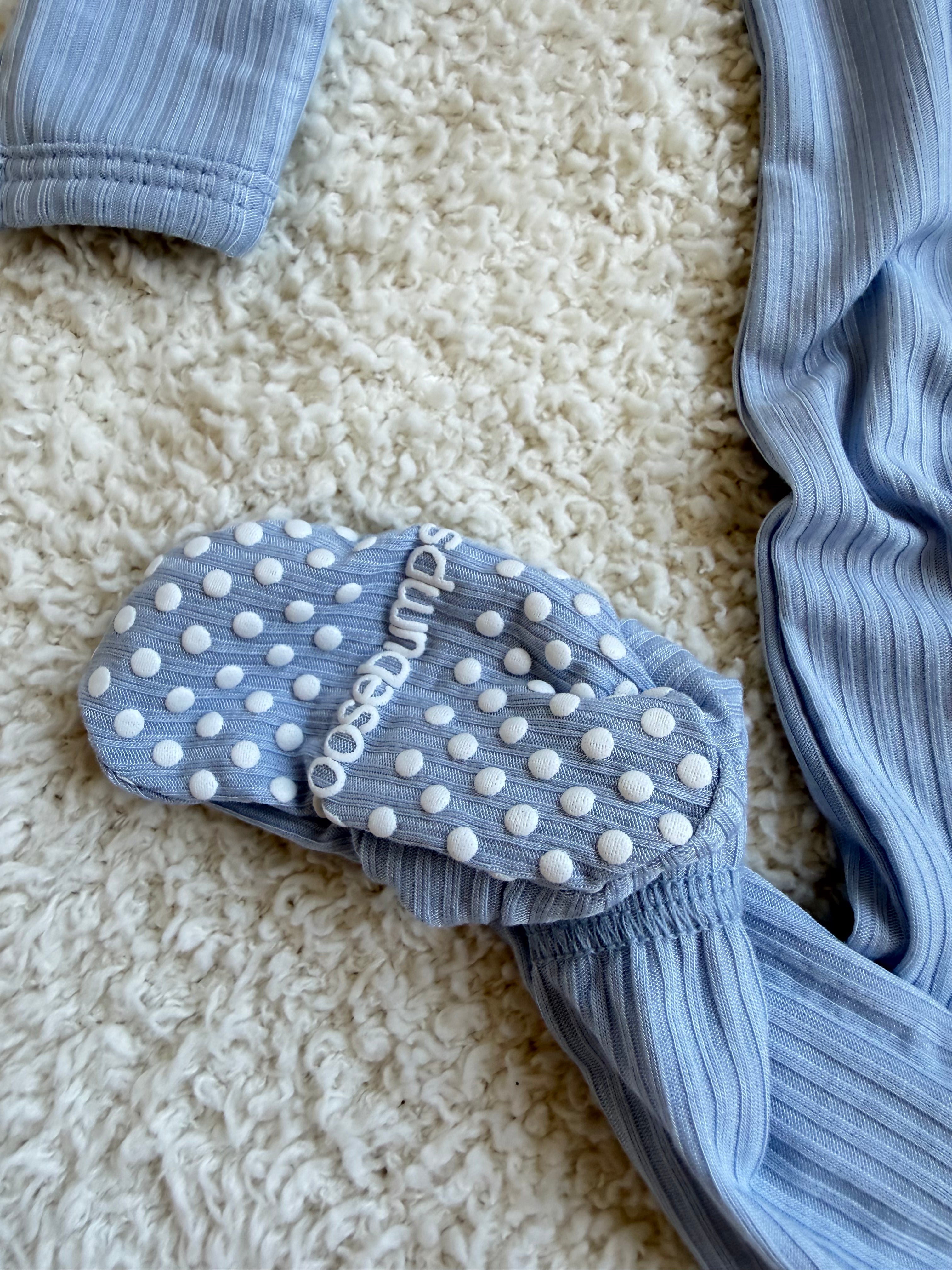 A cove / one piece zipper, light blue ribbed bamboo baby sleeper, rests on a cream textured carpet. One foot is visible with white non-slip dots and partial “dunsos” text on the sole.