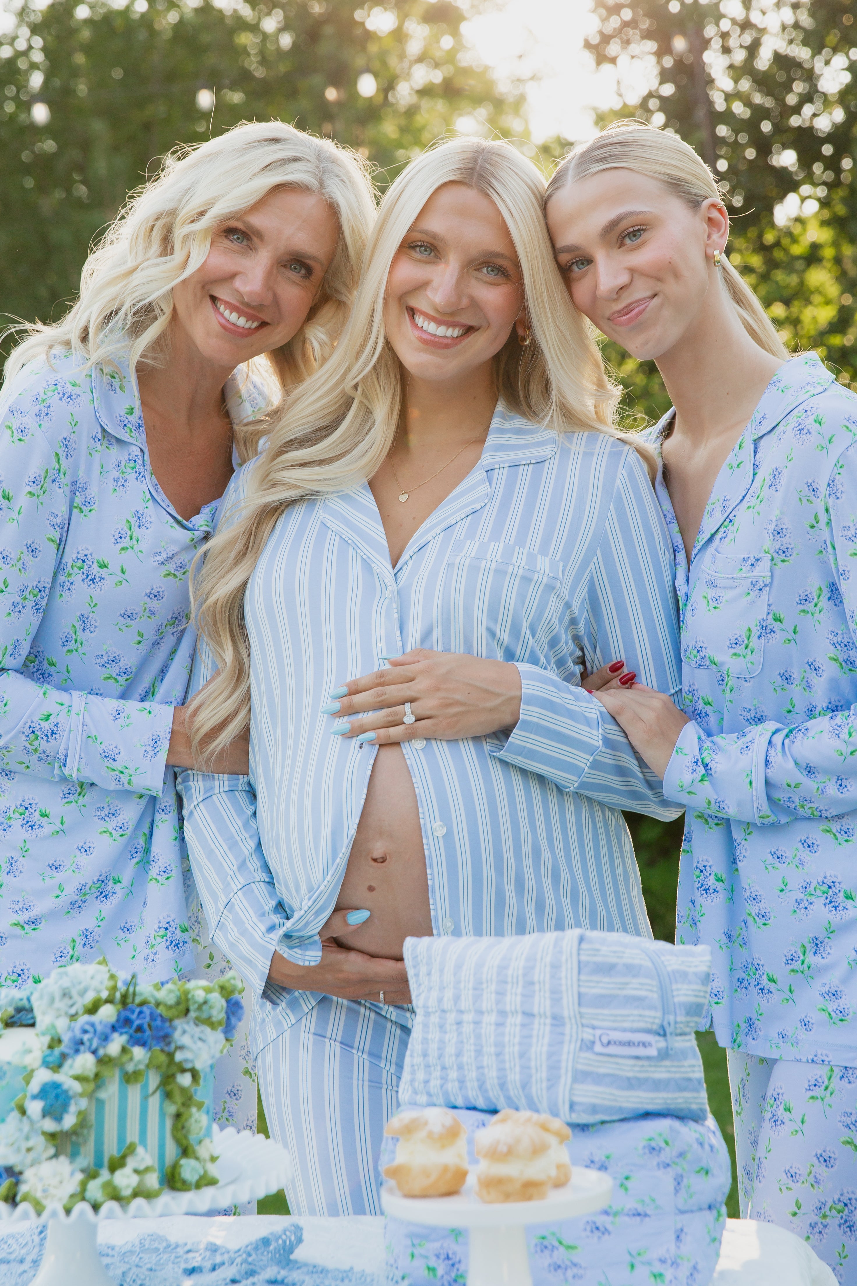Three blonde women in pastel blue pajamas stand outdoors, smiling. The center woman cradles her pregnant belly, decorated with blue hearts. They radiate comfort and joy clad in the haven pajama set (long pant). A cake, flowers, and scones sit on a table in the foreground.