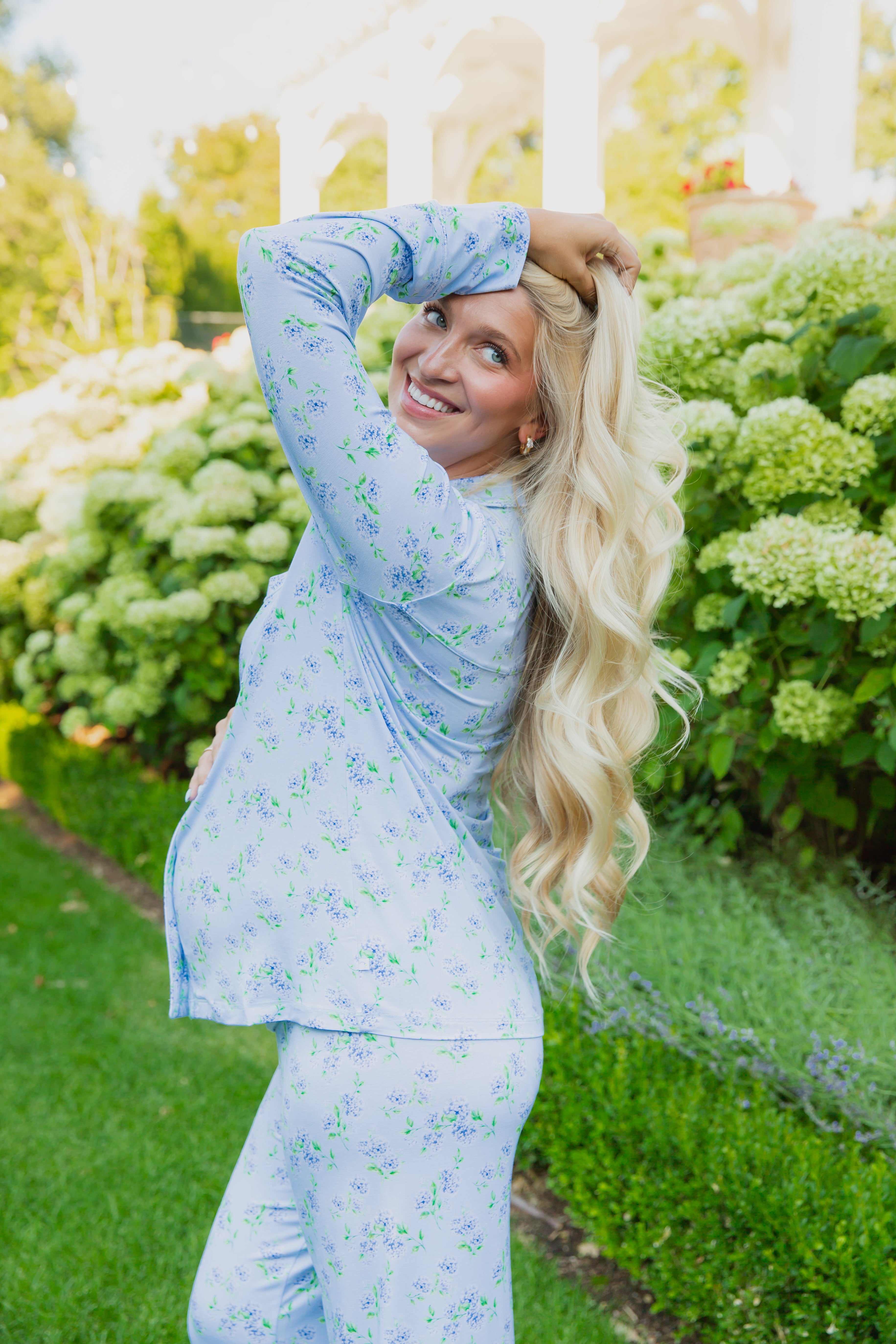 A smiling pregnant woman with long blonde hair poses outdoors among green bushes and white flowers, wearing the hydrangea pajama set with long pants from the Sarah Sweeten Goosebumps Collection.