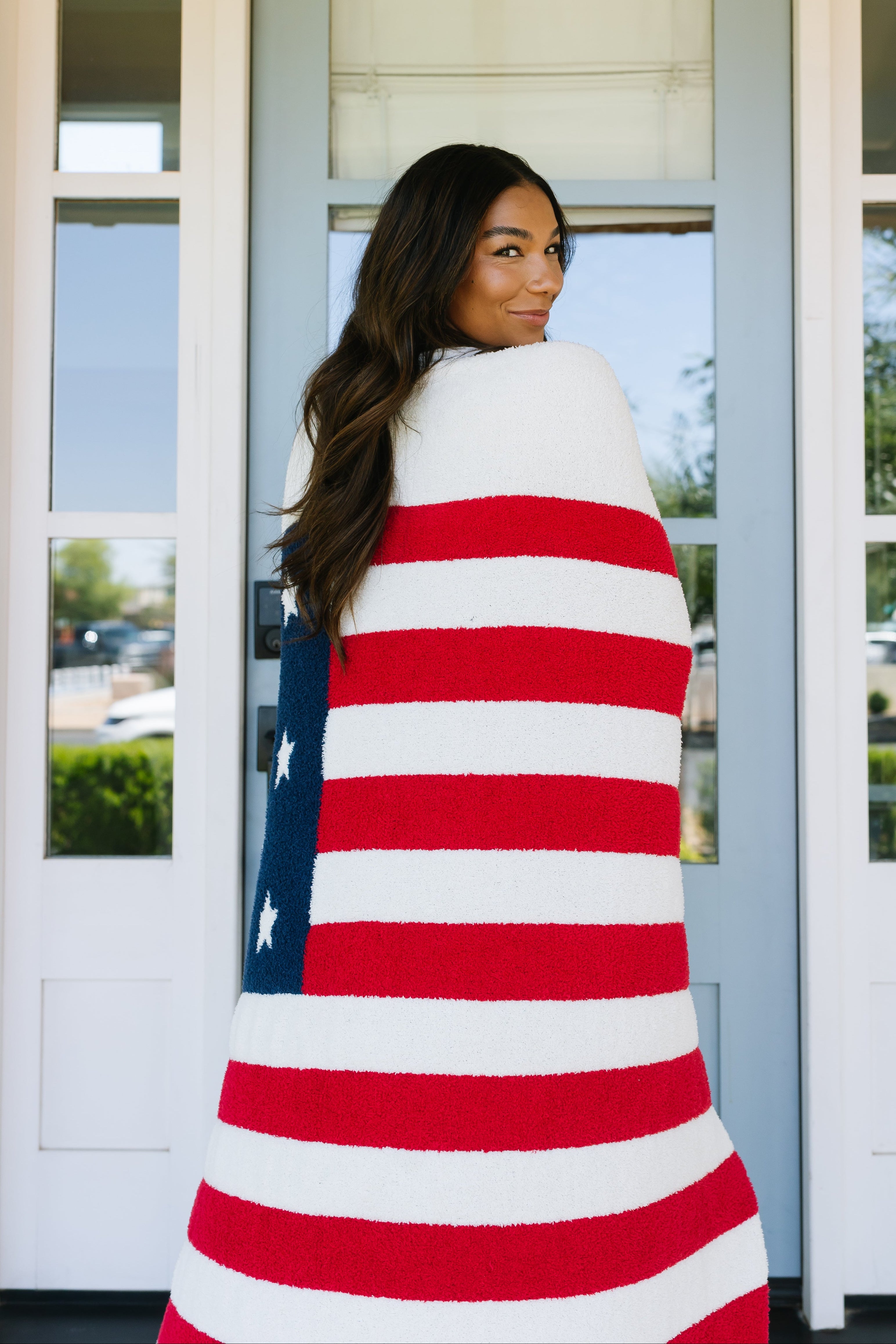 A woman with long dark hair smiles over her shoulder while wrapped in the america / plush blanket, standing before a pale blue door.