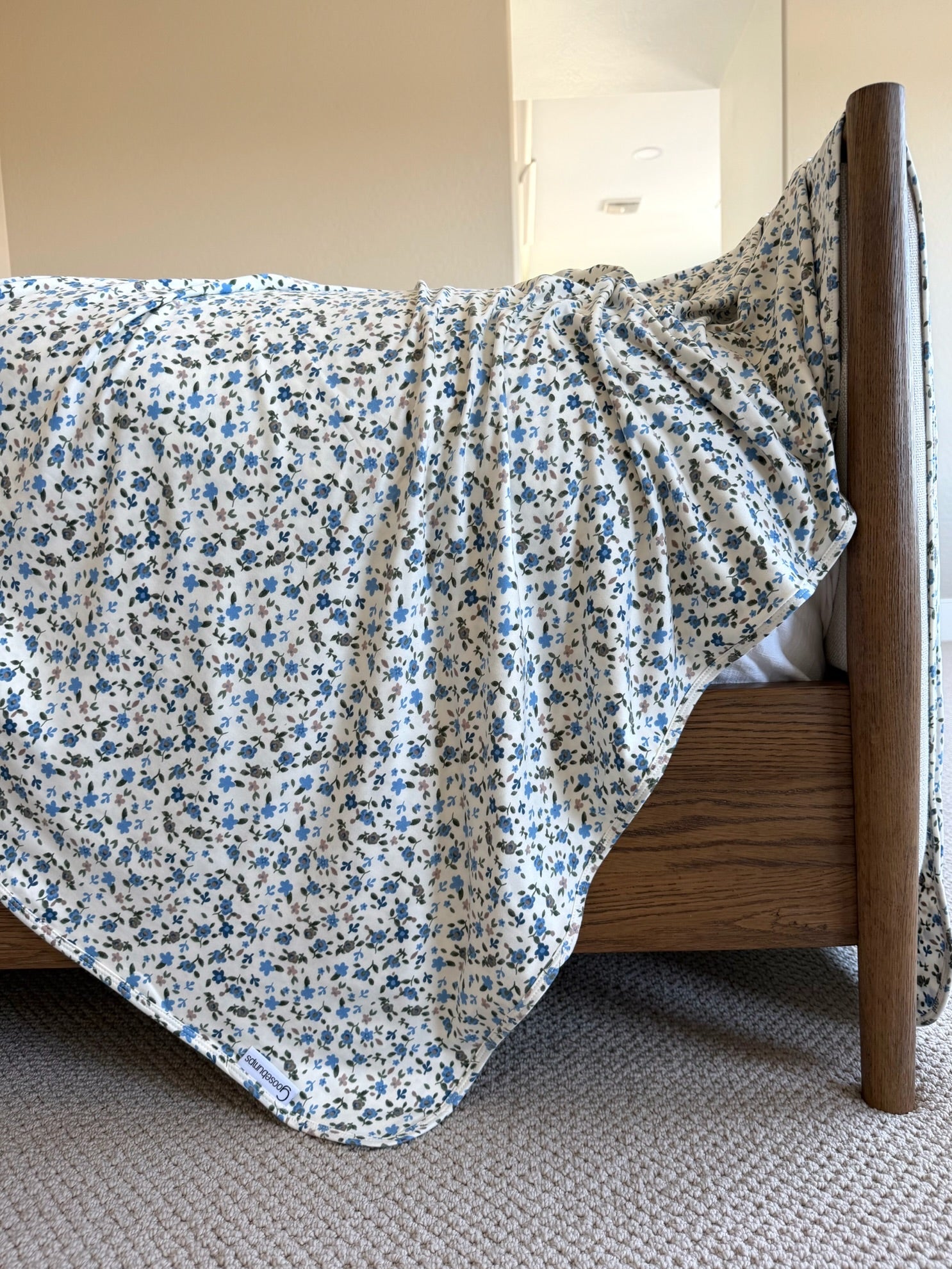 A wooden bed features the "choose me" blanket in soft blue and white floral patterns, elegantly draped over the side and touching a beige carpeted floor, while light streams in from a bright room beyond.