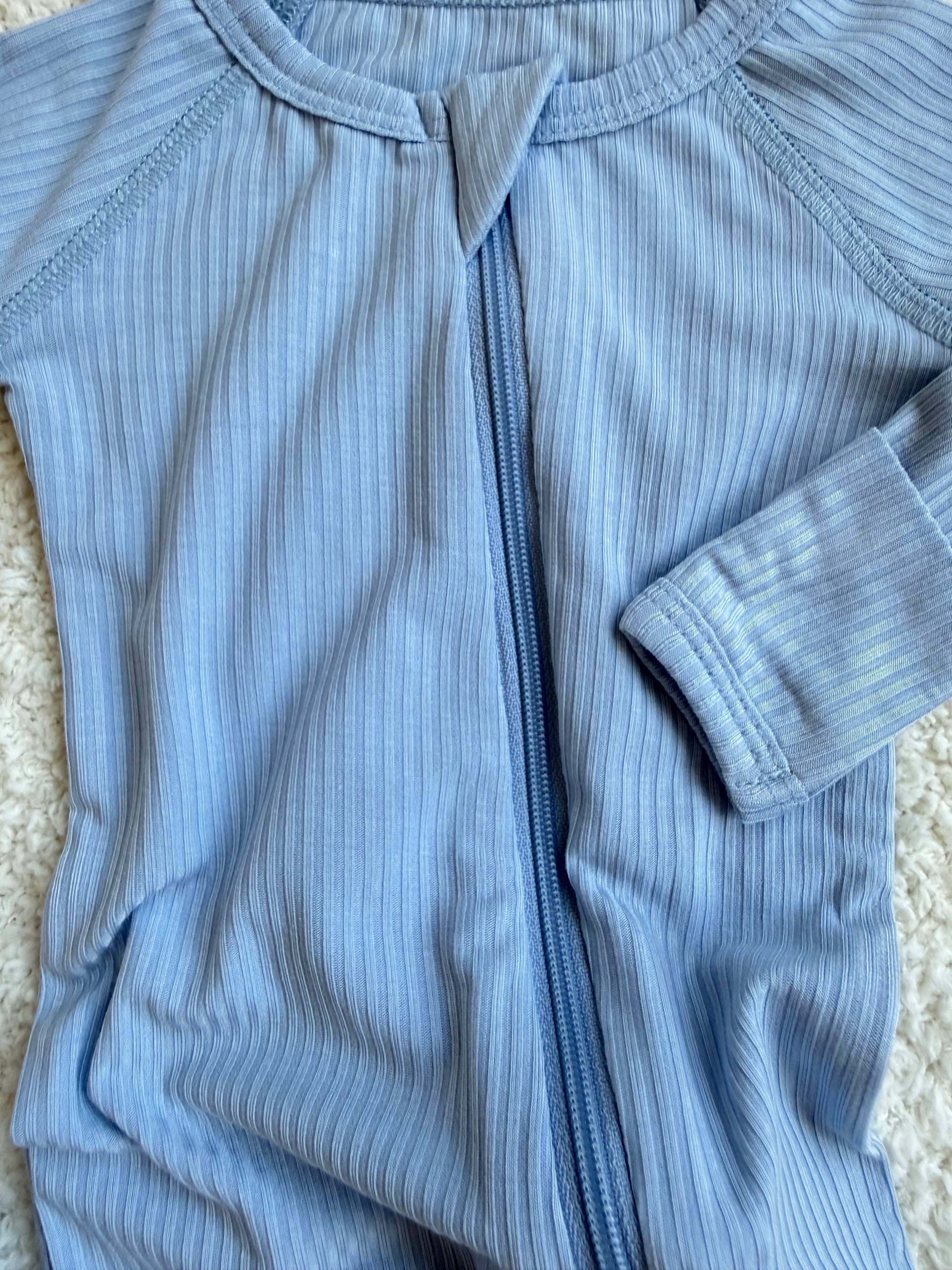 A close-up of the cove one piece zipper, a light blue, ribbed bamboo baby sleeper with a center zipper, displayed on a textured white surface.