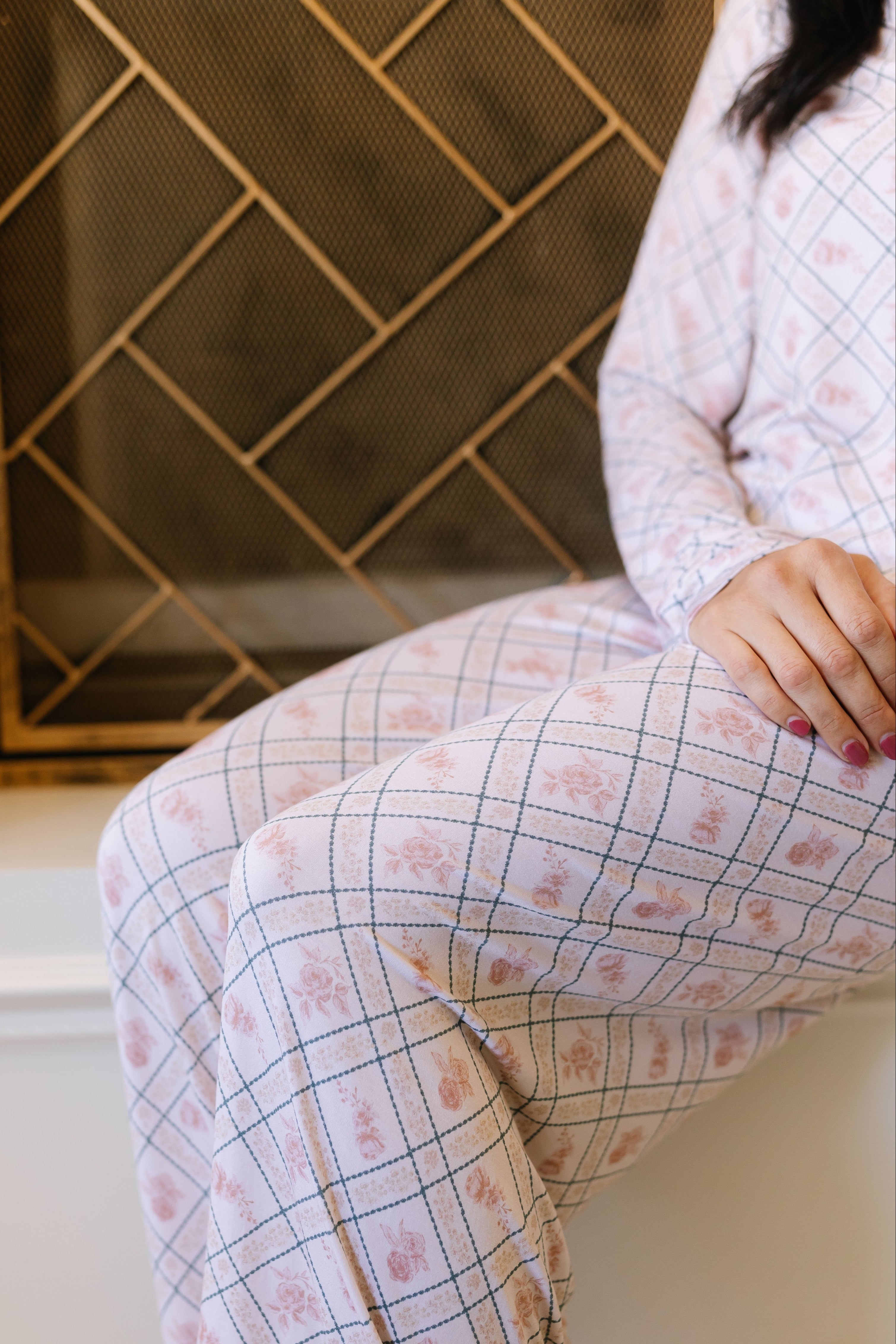 Wearing "the rose / pajama set," a person sits on a white surface near a decorative gold metal screen. Only their lower body and one arm are visible.