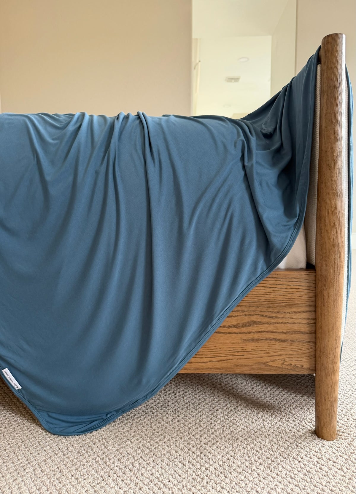A luxurious tidal wave blanket drapes over the side of a wooden bed frame in a carpeted bedroom, with a mirror and beige walls in the background.