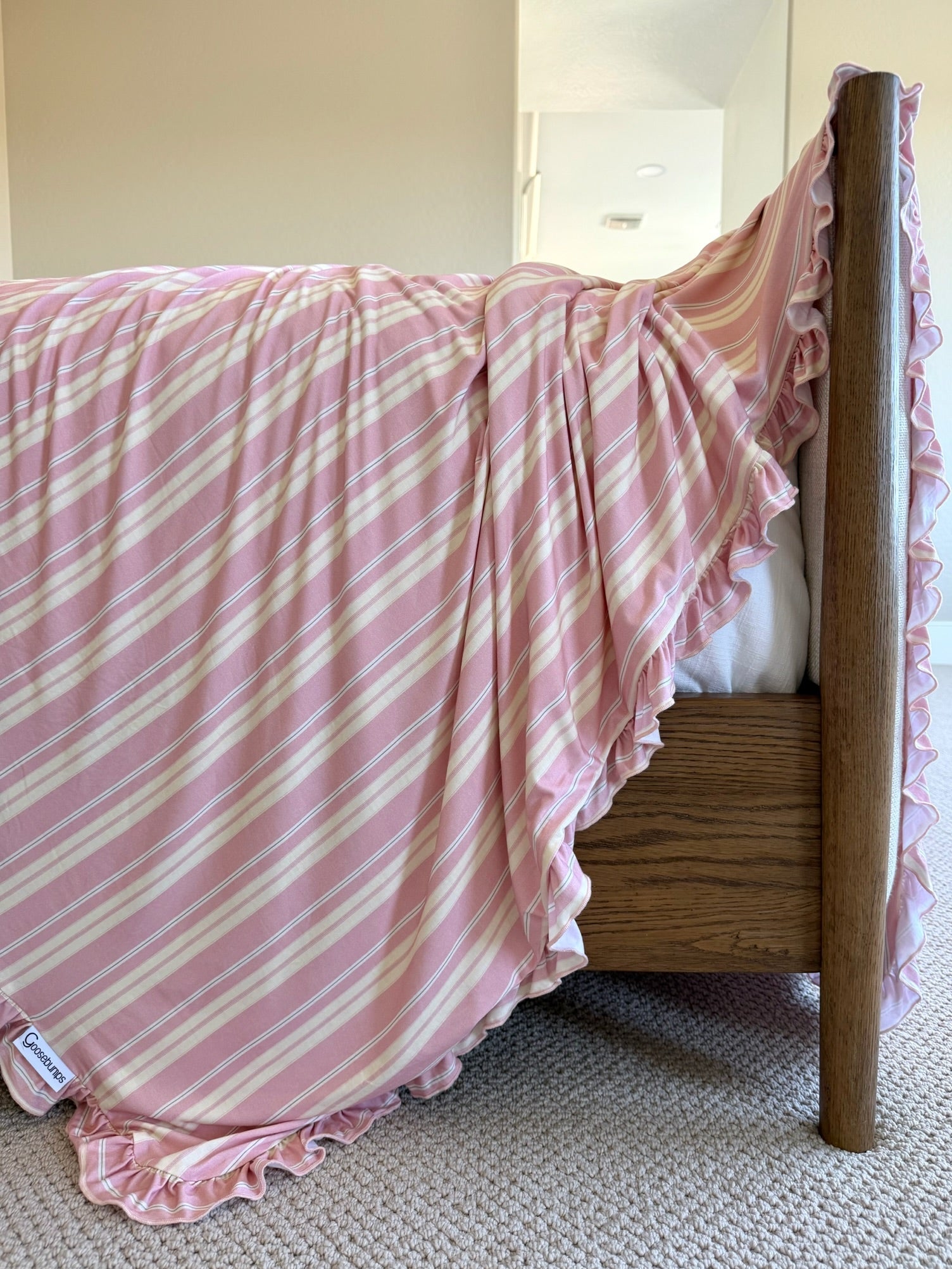 The sherbet / ruffle blanket, with pink and white stripes and elegant ruffles, drapes over a wooden bed and touches the beige carpet in a bright room.