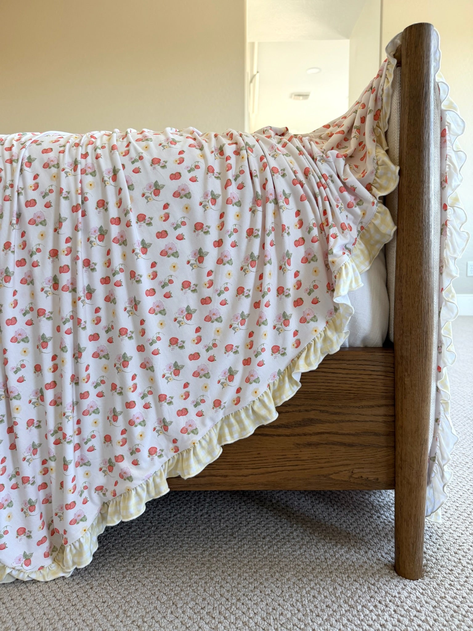 The freshly picked w/ sunshine ruffle blanket, featuring pink and white florals with ruffled yellow edges, is draped over a wooden bed on a carpeted floor in a softly lit bedroom.