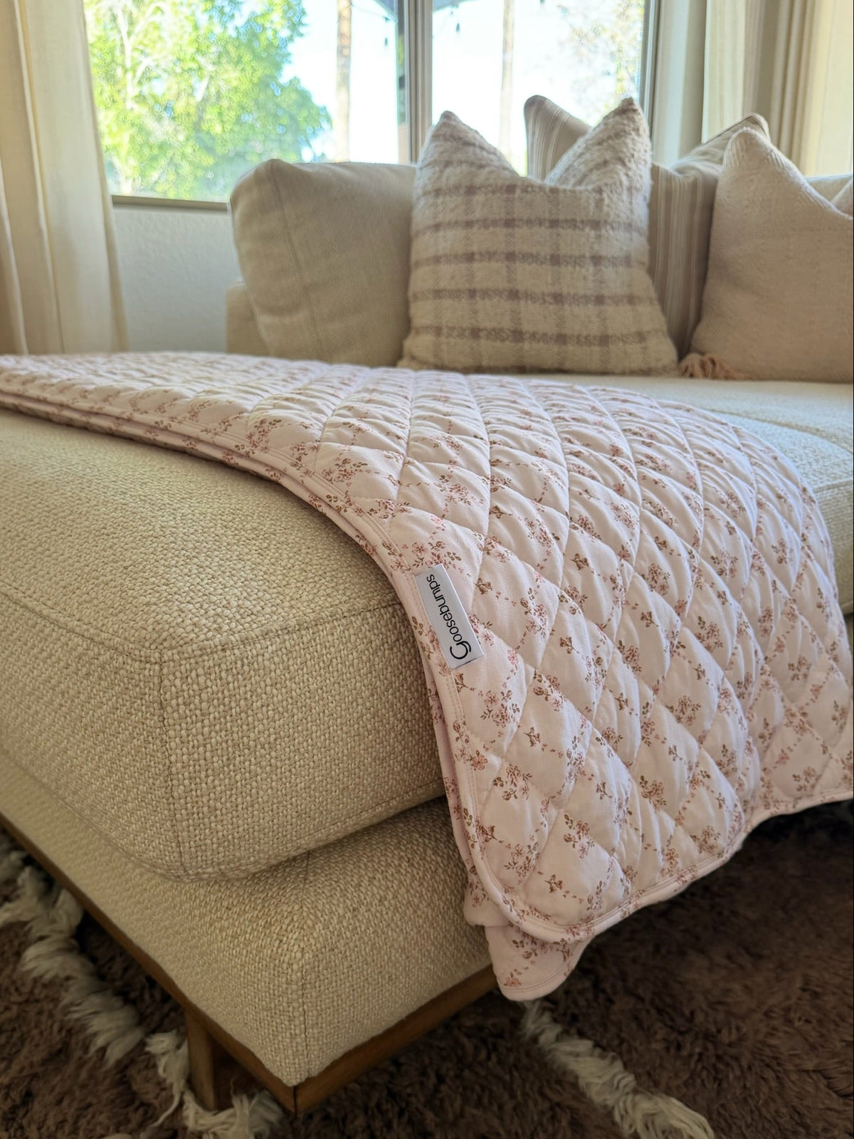 A keepsake / quilt in pink bamboo with a delicate floral print is draped over a beige textured couch adorned with plaid pillows, illuminated by sunlight streaming through the nearby window.