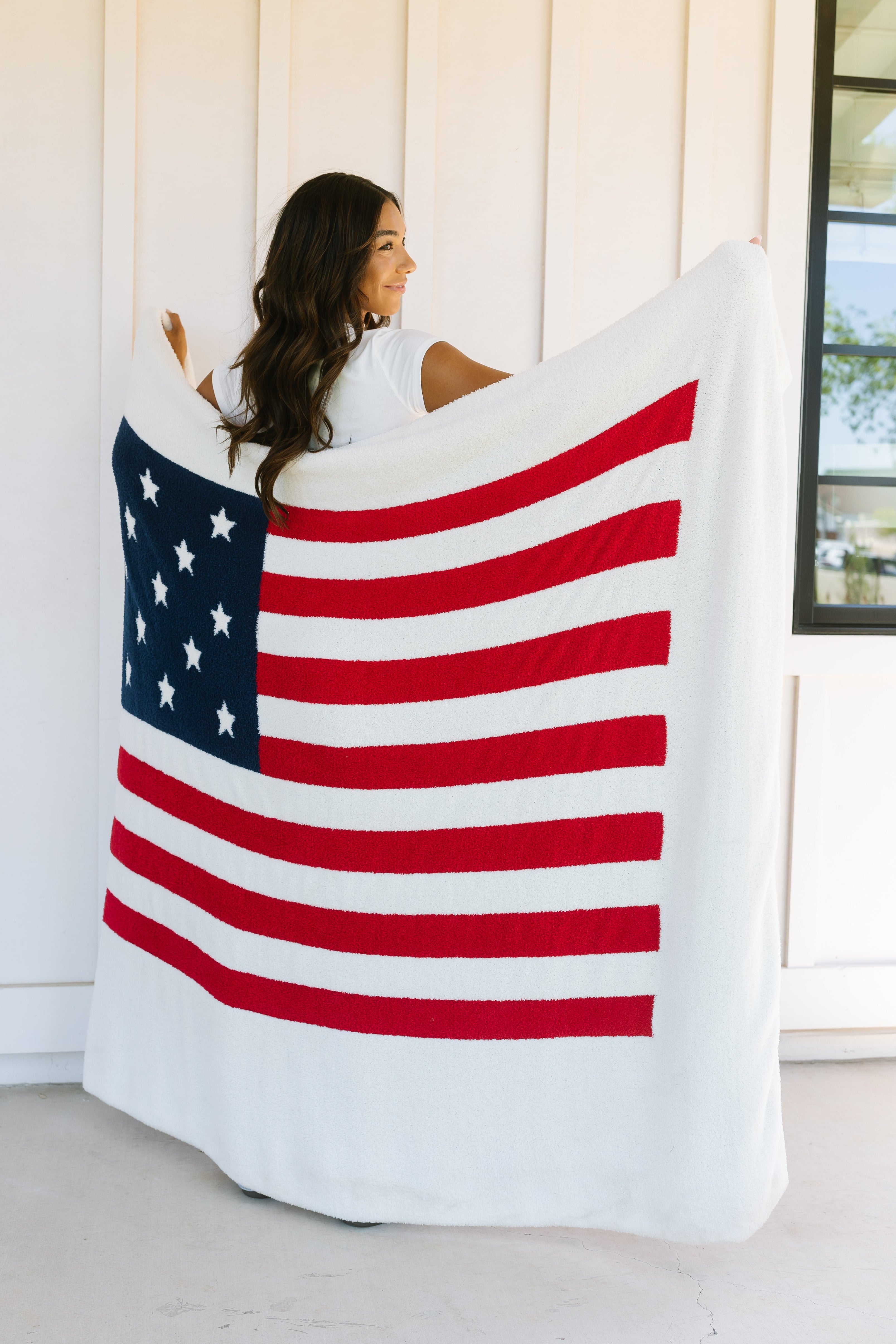 A woman smiles indoors, wrapped in the america / plush luxury lightweight blanket, with white walls and a window behind her.