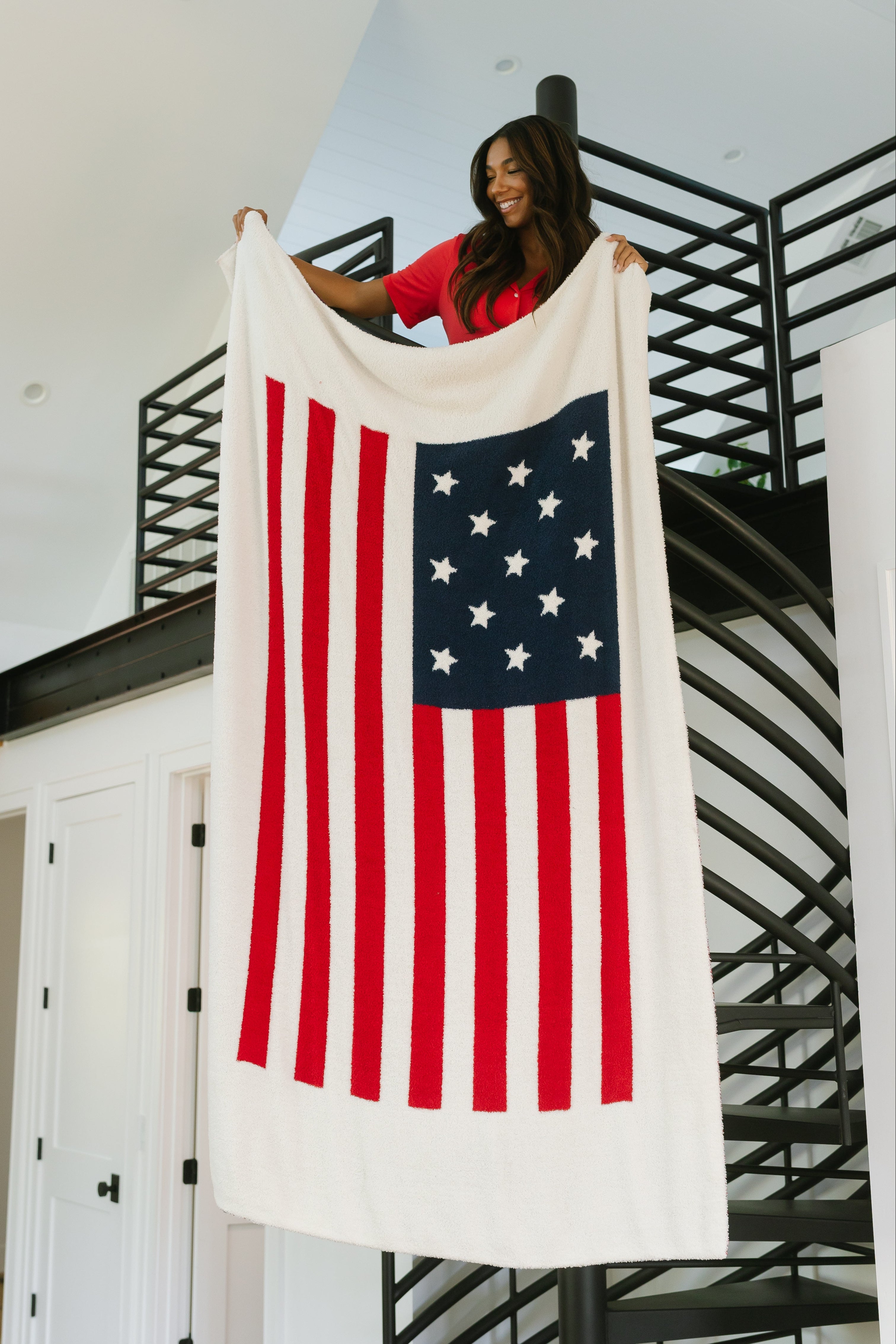 A woman stands on an indoor spiral staircase, holding the america / plush luxury lightweight blanket with red stripes and a central blue star square, adding a modern accent to the contemporary decor.