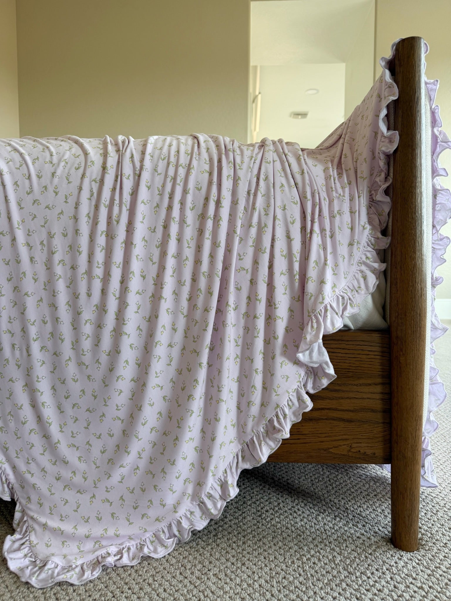 A wooden bed is draped with the rosebud / ruffle blanket in light purple, showcasing a delicate floral pattern and ruffled edges, flowing over the side onto a beige carpet, set against neutral walls.