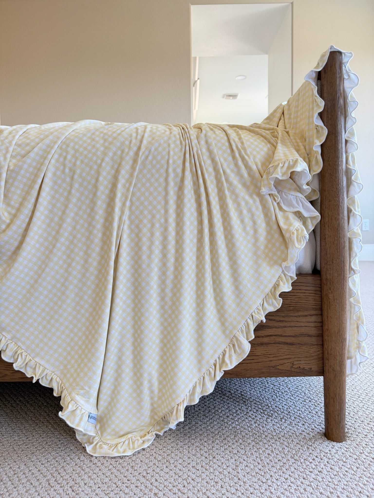 The sunshine / ruffle blanket, featuring light yellow and white checks with elegant ruffled edges, drapes over a wooden bed frame in a bright, carpeted bedroom.