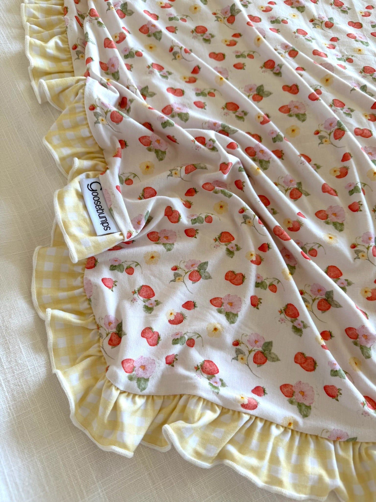 The "freshly picked w/ sunshine ruffle" bamboo blanket features red strawberries, white flowers, and a yellow-and-white gingham ruffle. A Goosebumps tag is attached, and the blanket is displayed on a white surface.