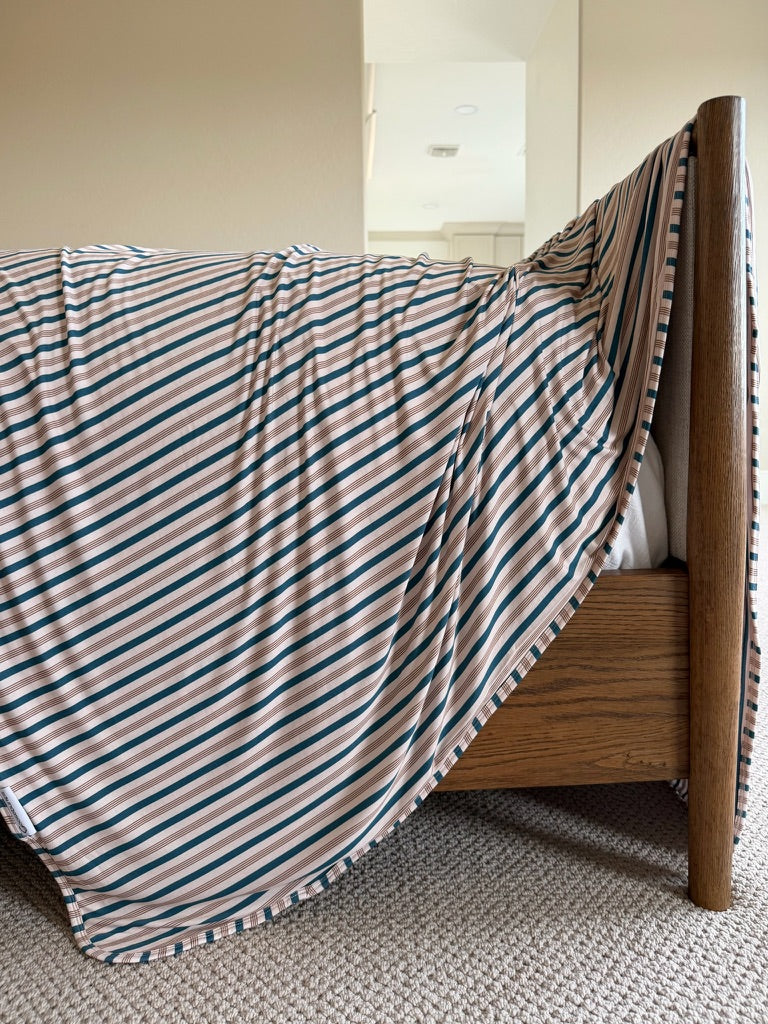 The Shoreline blanket, featuring elegant teal and beige stripes, is draped over the side of a wooden bed frame in a carpeted bedroom with neutral walls and a mirrored closet in the background.