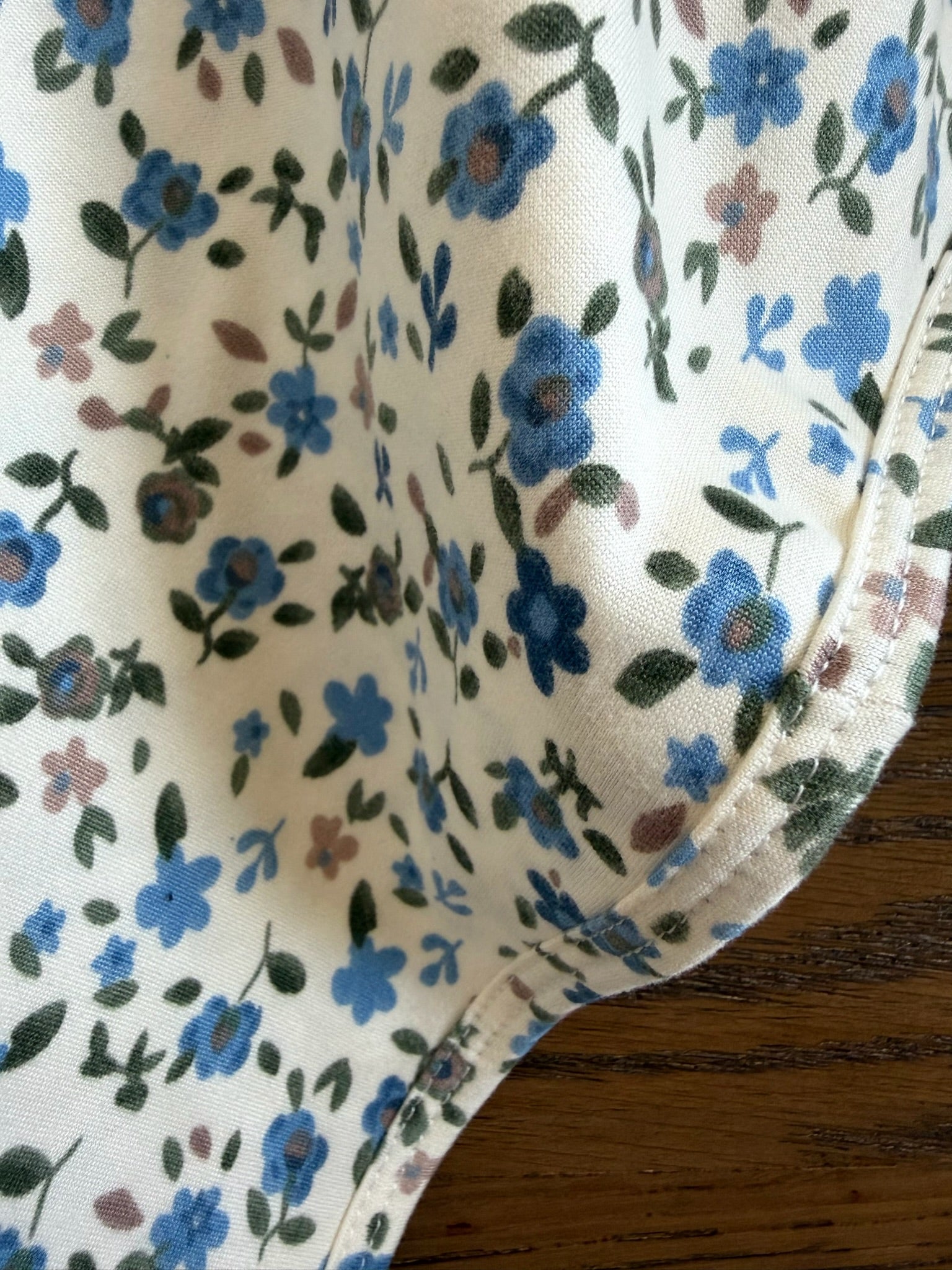 Close-up of "choose me," a cream-colored blanket featuring a blue and brown floral pattern and a visible stitched seam, gracefully draped on a wooden surface.