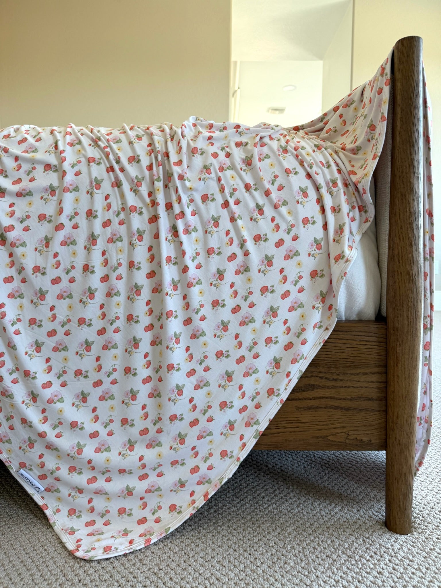 A wooden bed is adorned with the "freshly picked" blanket, featuring small red and green strawberry prints, in a carpeted room with neutral-colored walls.