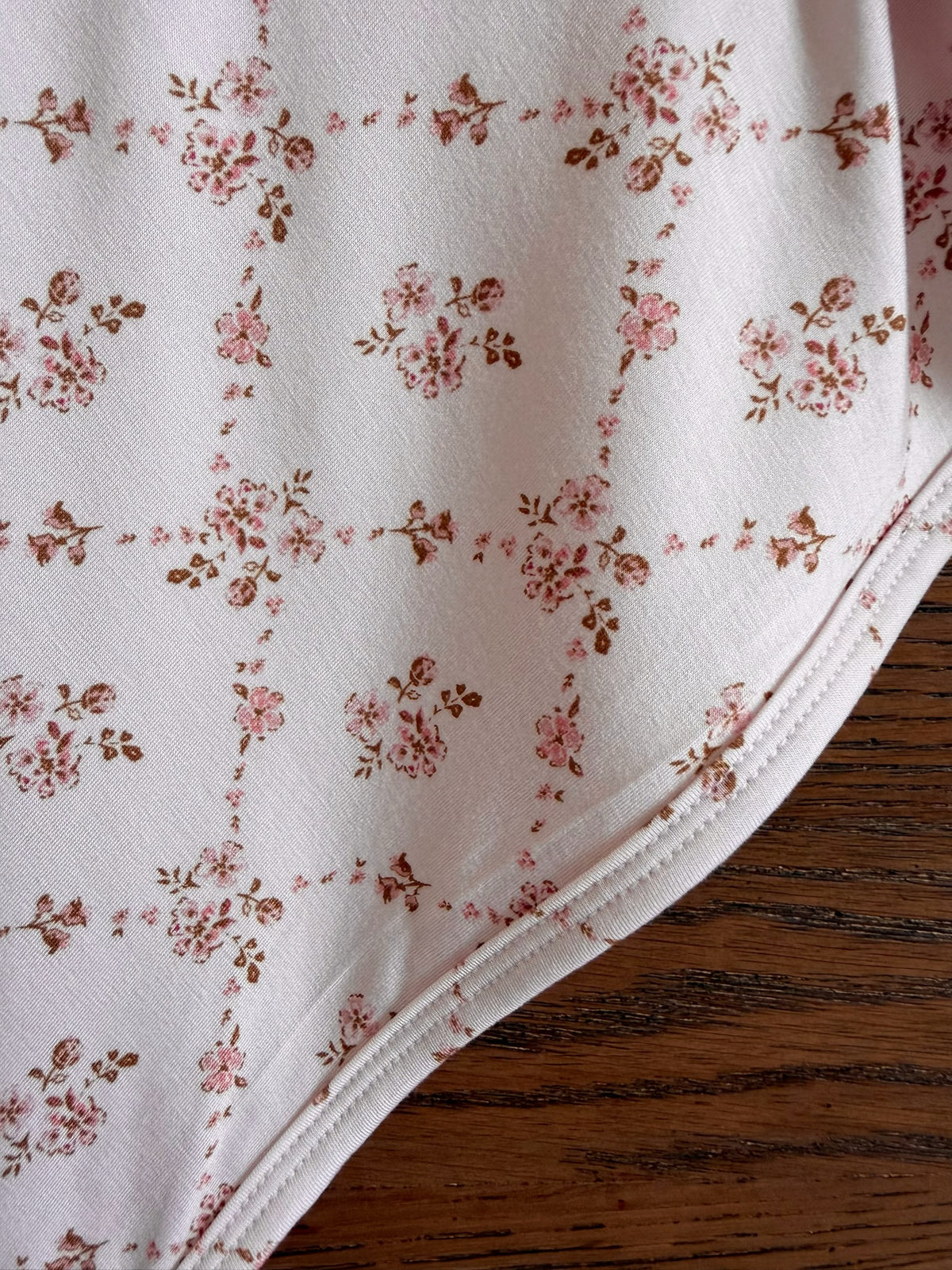 Close-up of the keepsake blanket, showcasing its luxurious white fabric adorned with a delicate pink and brown floral grid pattern. The blanket’s edges are elegantly curved and finely stitched, reflecting exceptional craftsmanship. A wooden surface peeks out from beneath, enhancing the refined details of this floral print keepsake blanket.