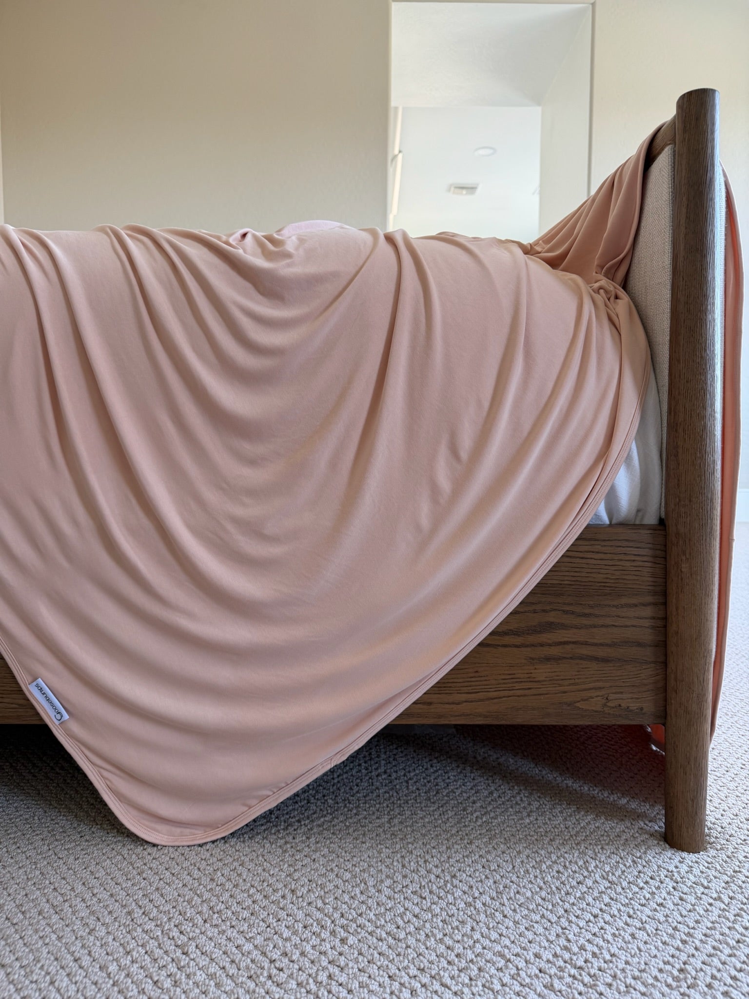 A whisp adult swaddle blanket in light pink is draped over the side of a wooden bed, lying smoothly with gentle folds on beige carpeting. A white tag is visible on one corner of the blanket.