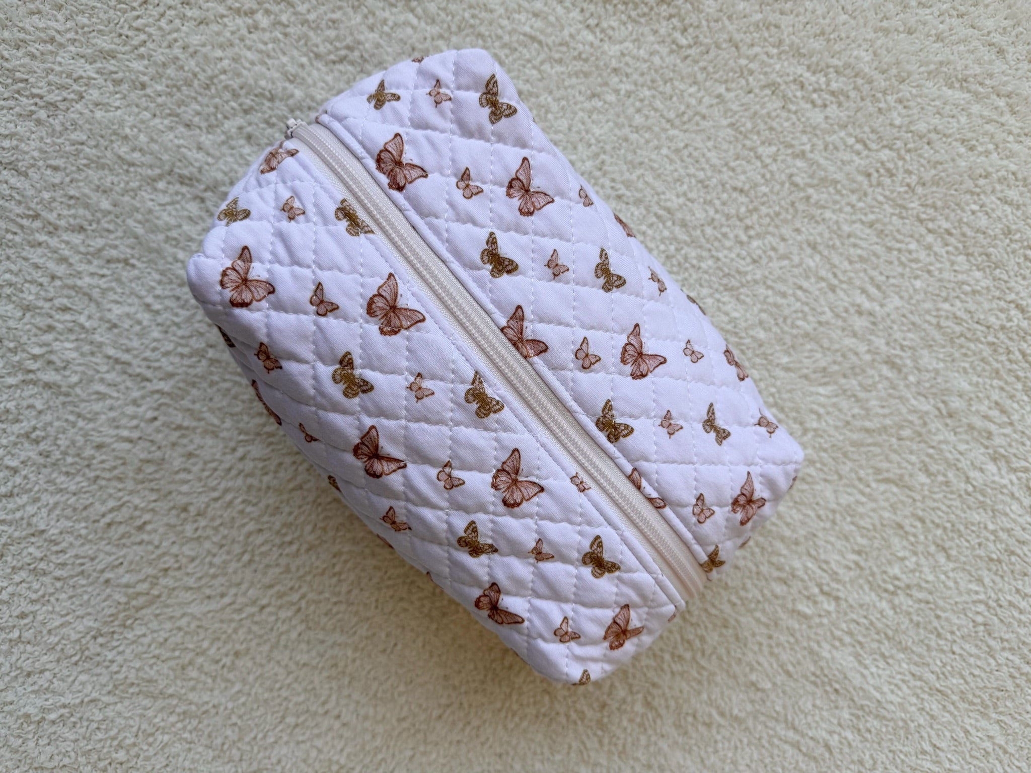 The flutter / travel bag is a quilted, rectangular zipper bag with a feminine butterfly print on light beige bamboo viscose, displayed on a textured, light surface.