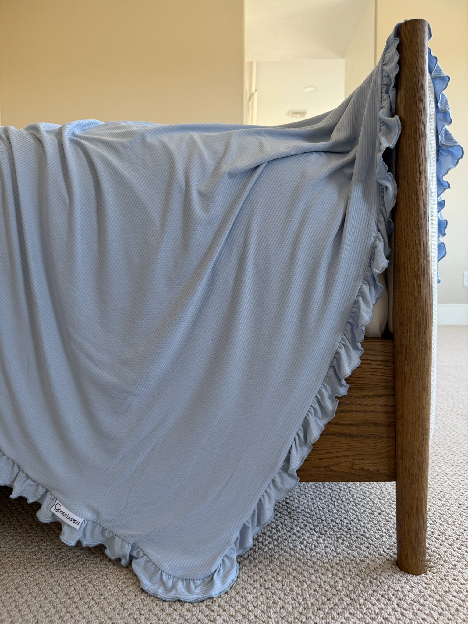 The cove ribbed ruffle blanket in light blue drapes over a wooden bed frame in a sunlit room with beige carpet and neutral walls, its ruffled edges and small white tag adding a touch of luxury.