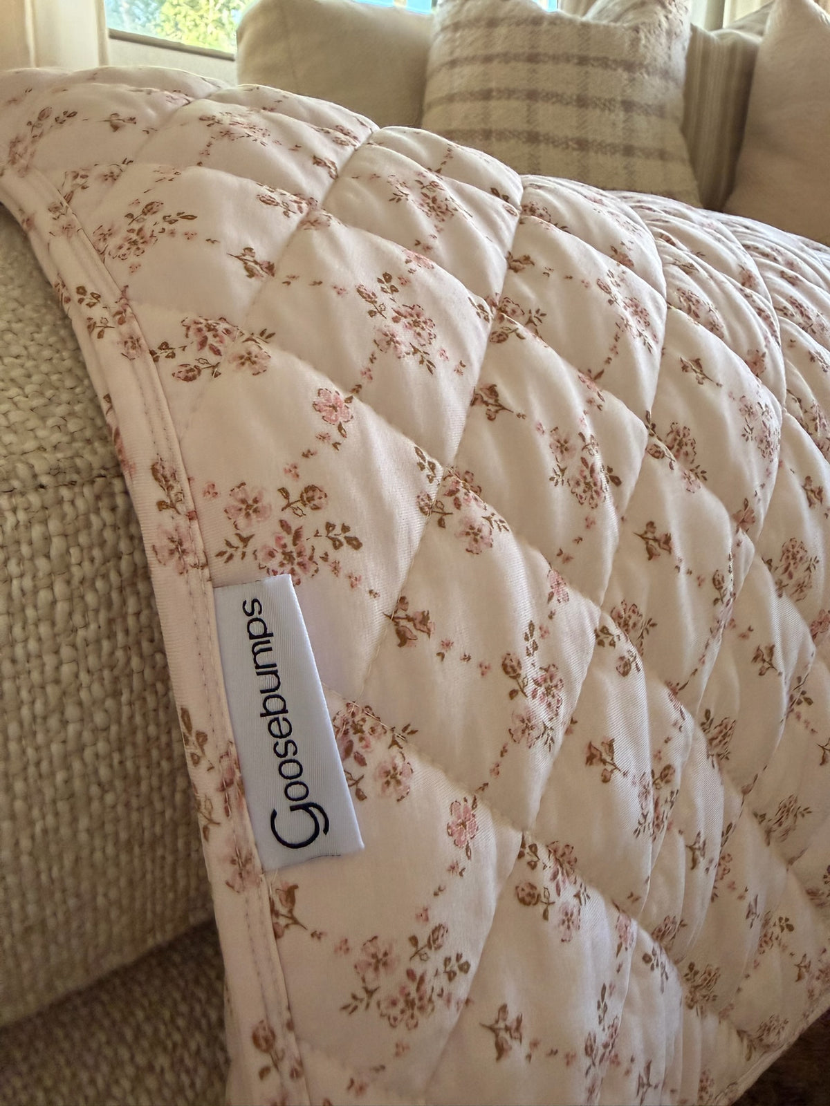A keepsake / quilt featuring a delicate pink floral print and a Goosebumps label is draped over a beige textured sofa, with pillows in the background—perfect for creating a cozy adult swaddle experience.