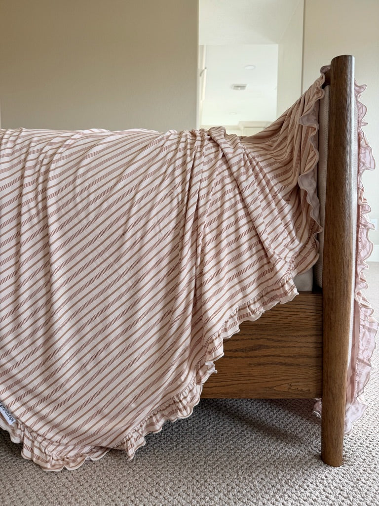 A wooden bed with the toast and honey / ruffle blanket draped over the side, set on a light carpeted floor in a softly lit room.