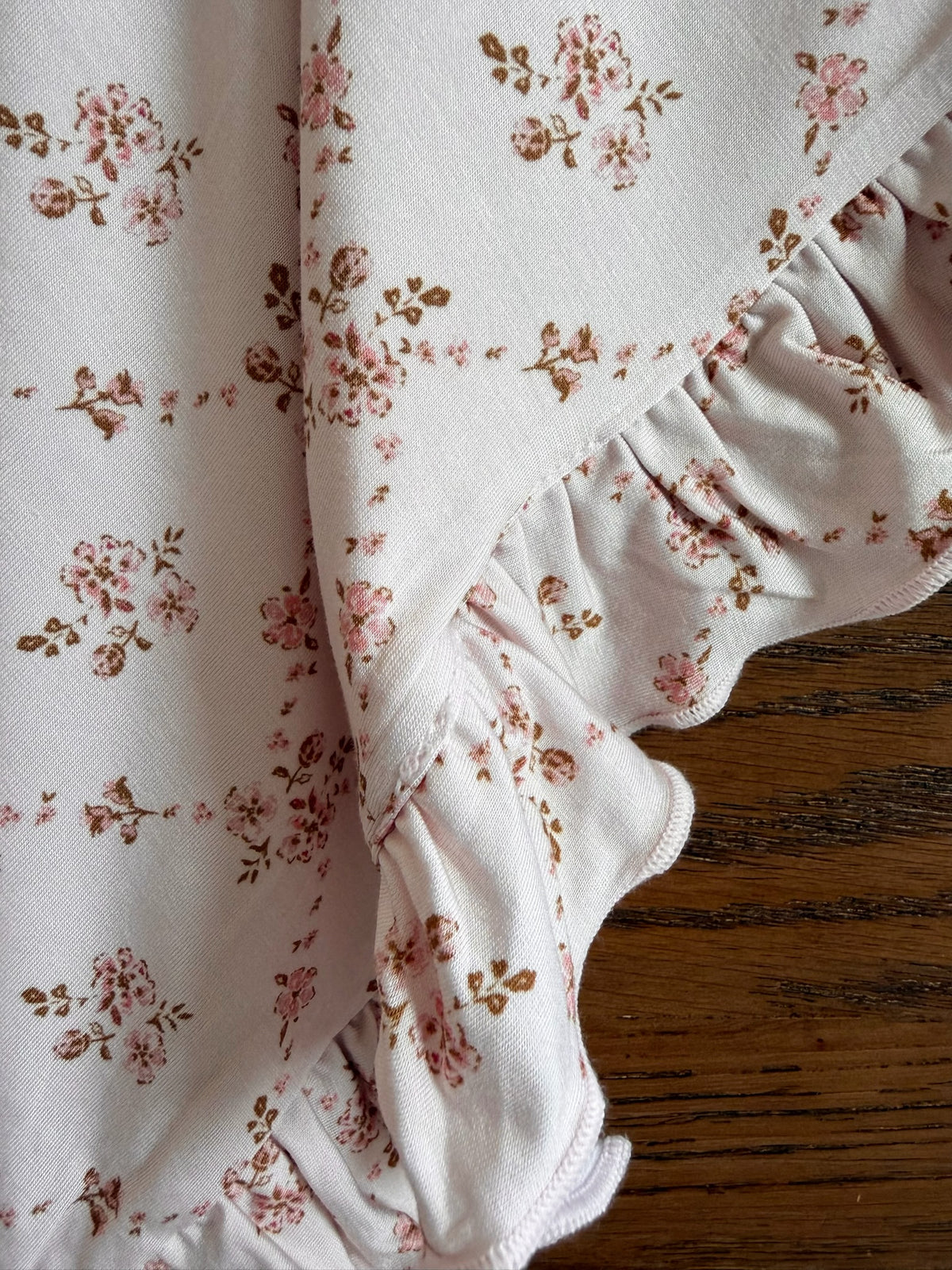 A close-up of the keepsake / ruffle, featuring white ruffled fabric adorned with a small pink and brown floral pattern, rests on a wooden surface—perfectly capturing the delicate beauty of this floral blanket.