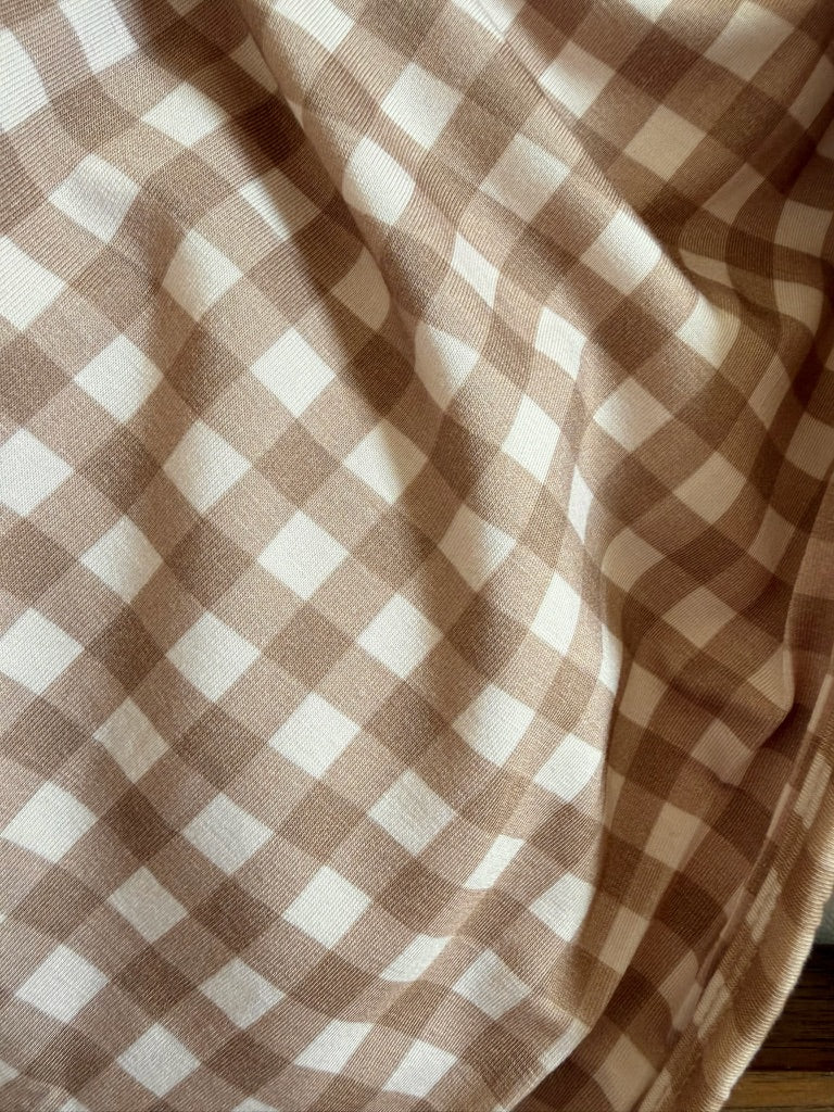 A close-up of tan and white checkered fabric in the signature teddy bear check pattern with gentle folds casting soft shadows—perfect as a luxurious blanket for cozy evenings or as a stylish travel companion on your next adventure.