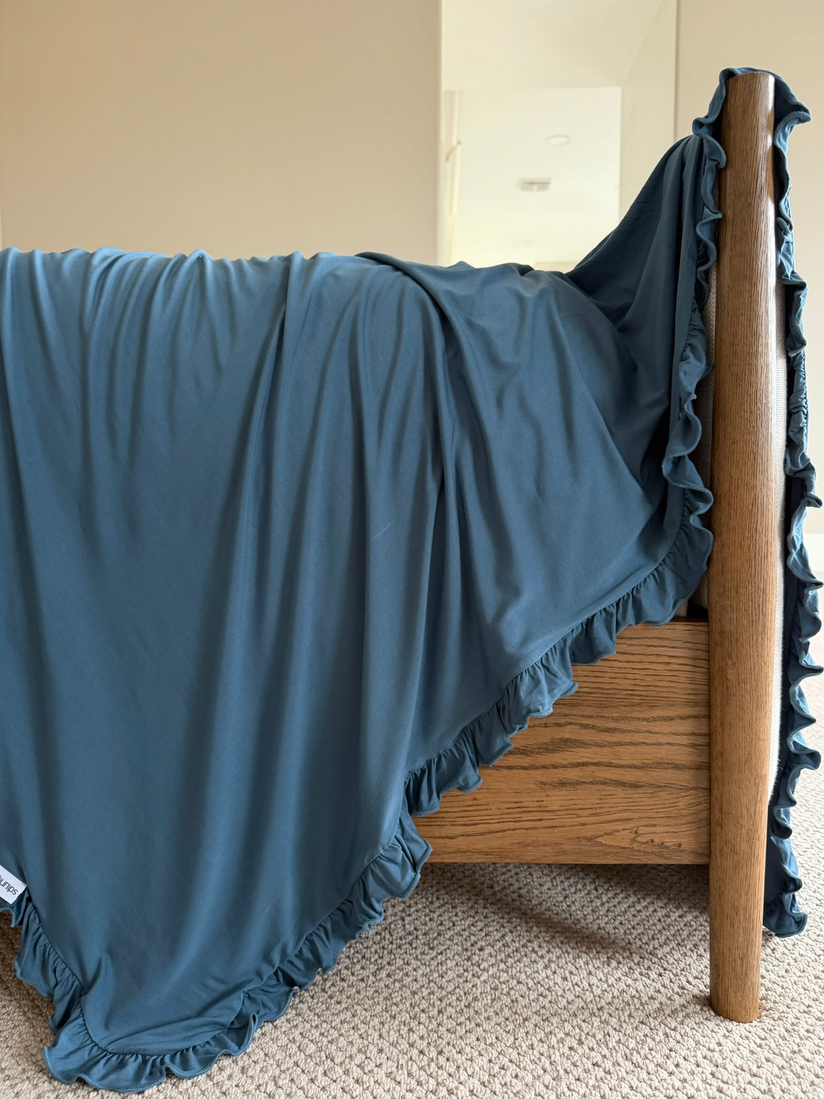 A wooden bed featuring the “tidal wave / ruffle” blue luxurious blanket draped over the side, set on a beige carpeted floor in a softly lit room.