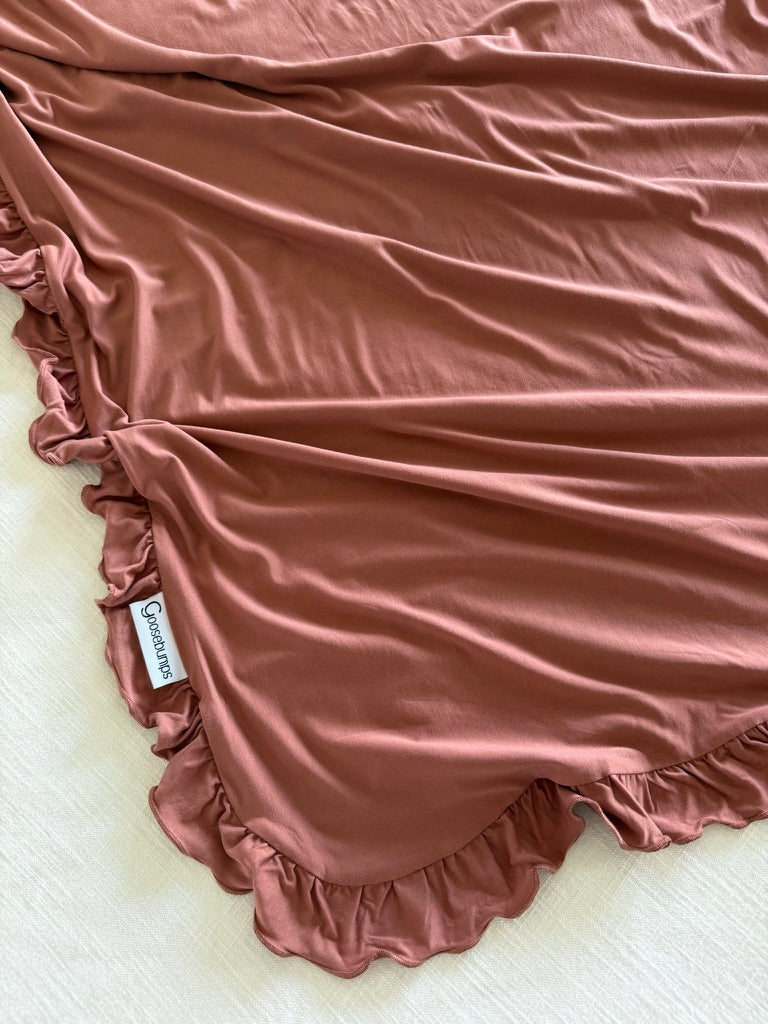A serenity / ruffle blanket in a soft, dusty rose hue with elegant ruffled edges is spread across a light surface. The edge is accented by a small white tag with black text.