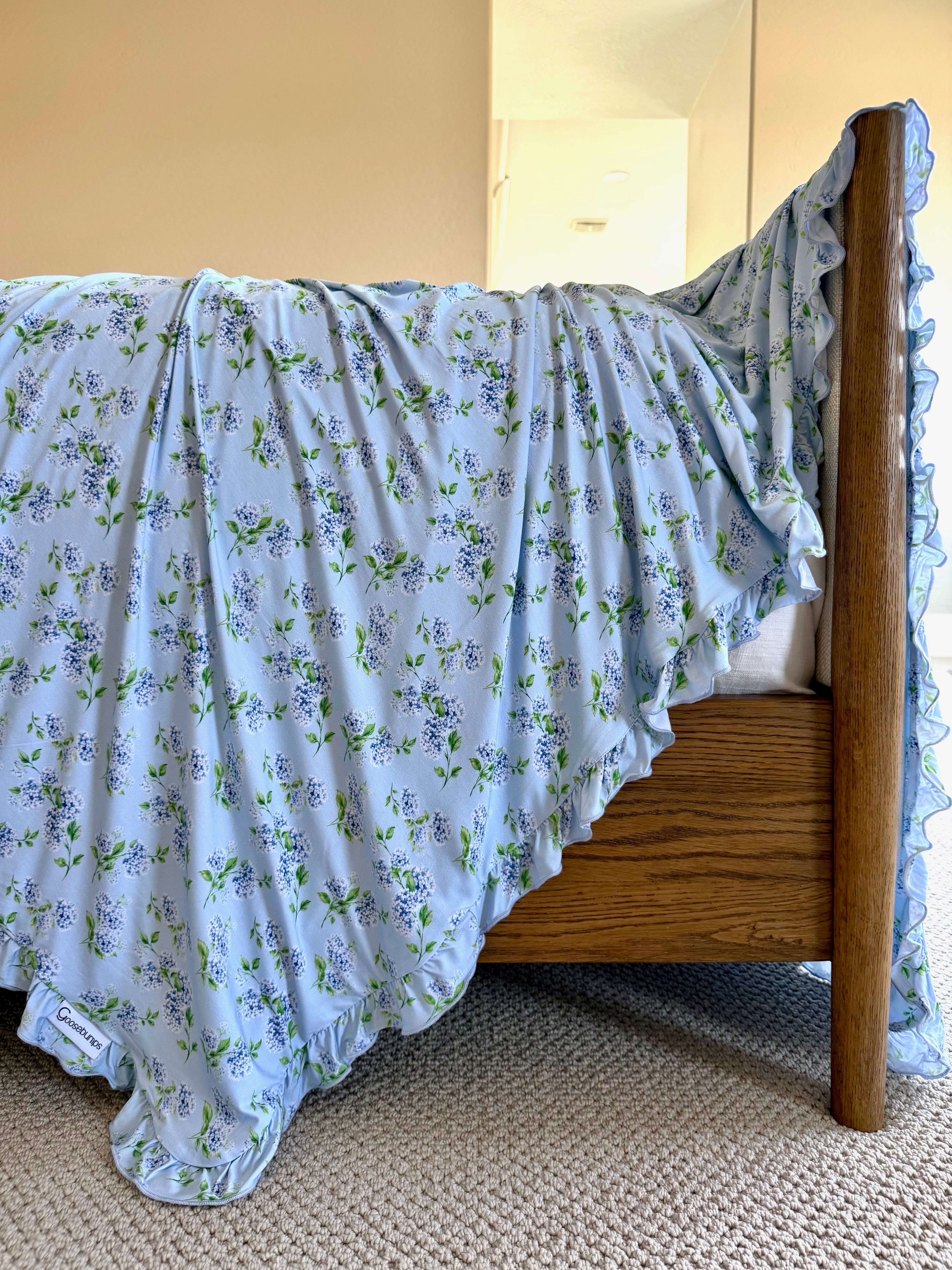 The hydrangea / ruffle blanket, featuring a light blue floral print and ruffled edges, is draped over the wooden footboard of a bed on a beige carpeted floor.