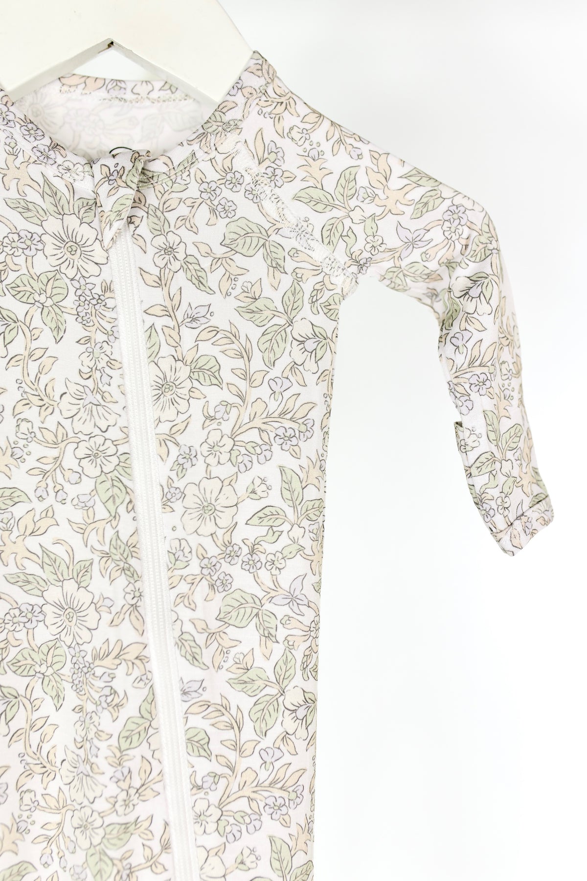A close-up of the heirloom / one piece zipper, a bamboo newborn sleeper with a floral pattern in soft green and beige tones, featuring a white two-way zipper down the front and long sleeves, hanging on a white hanger.