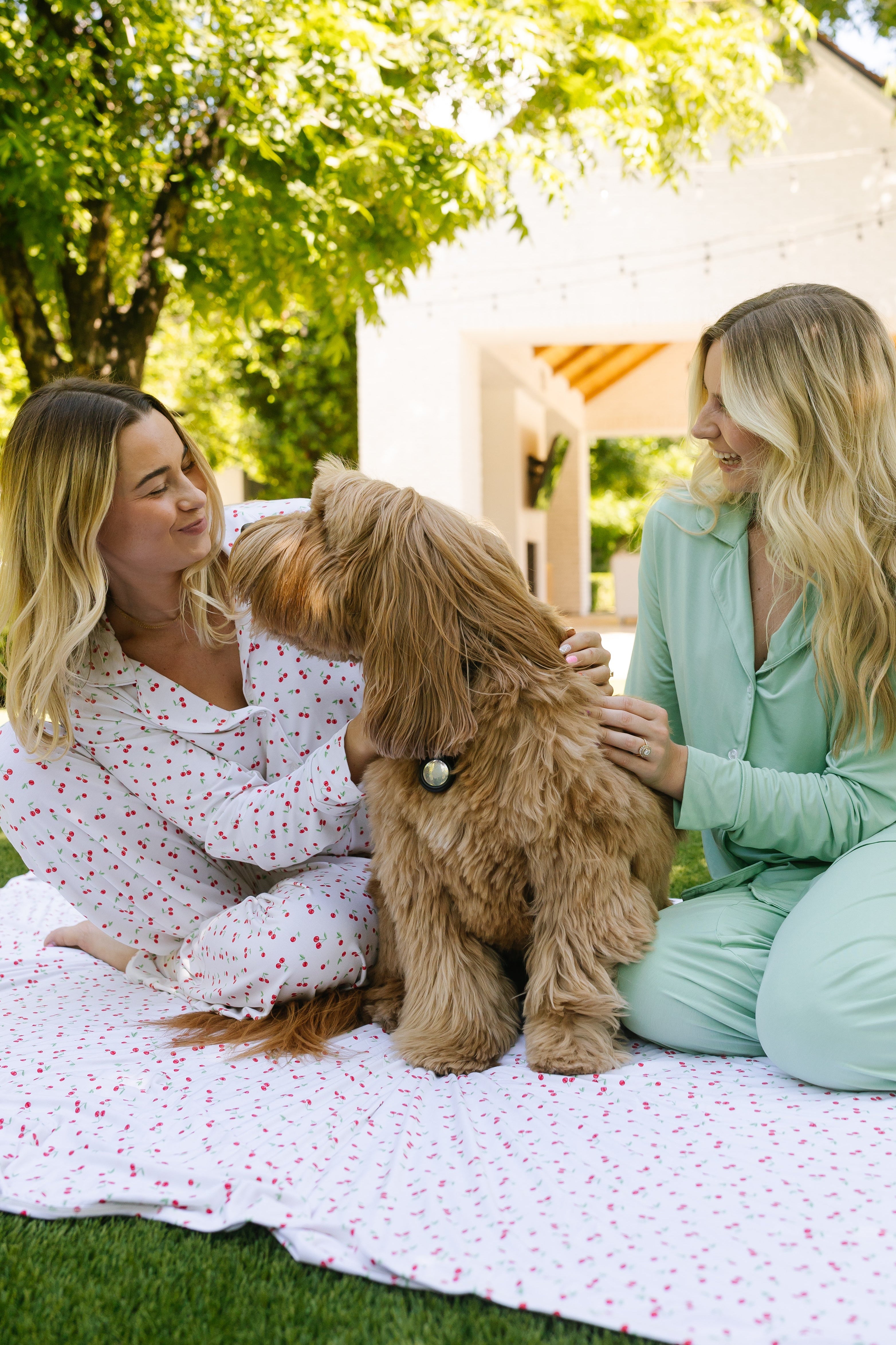 Two women in pajamas enjoy a sunny day outside, sitting on a luxurious cherry on top blanket in a grassy yard. They smile and pet a fluffy brown dog, with trees and a white house in the background.