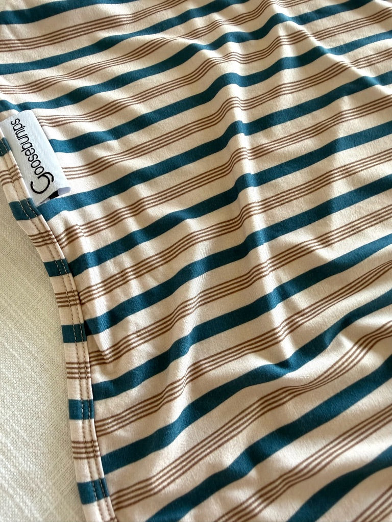A close-up of the Shoreline blanket—a soft, striped bamboo throw featuring alternating teal and brown lines, accented by a sewn white Cosebunny tag on the edge. The luxurious fabric is slightly wrinkled and draped over a light surface.