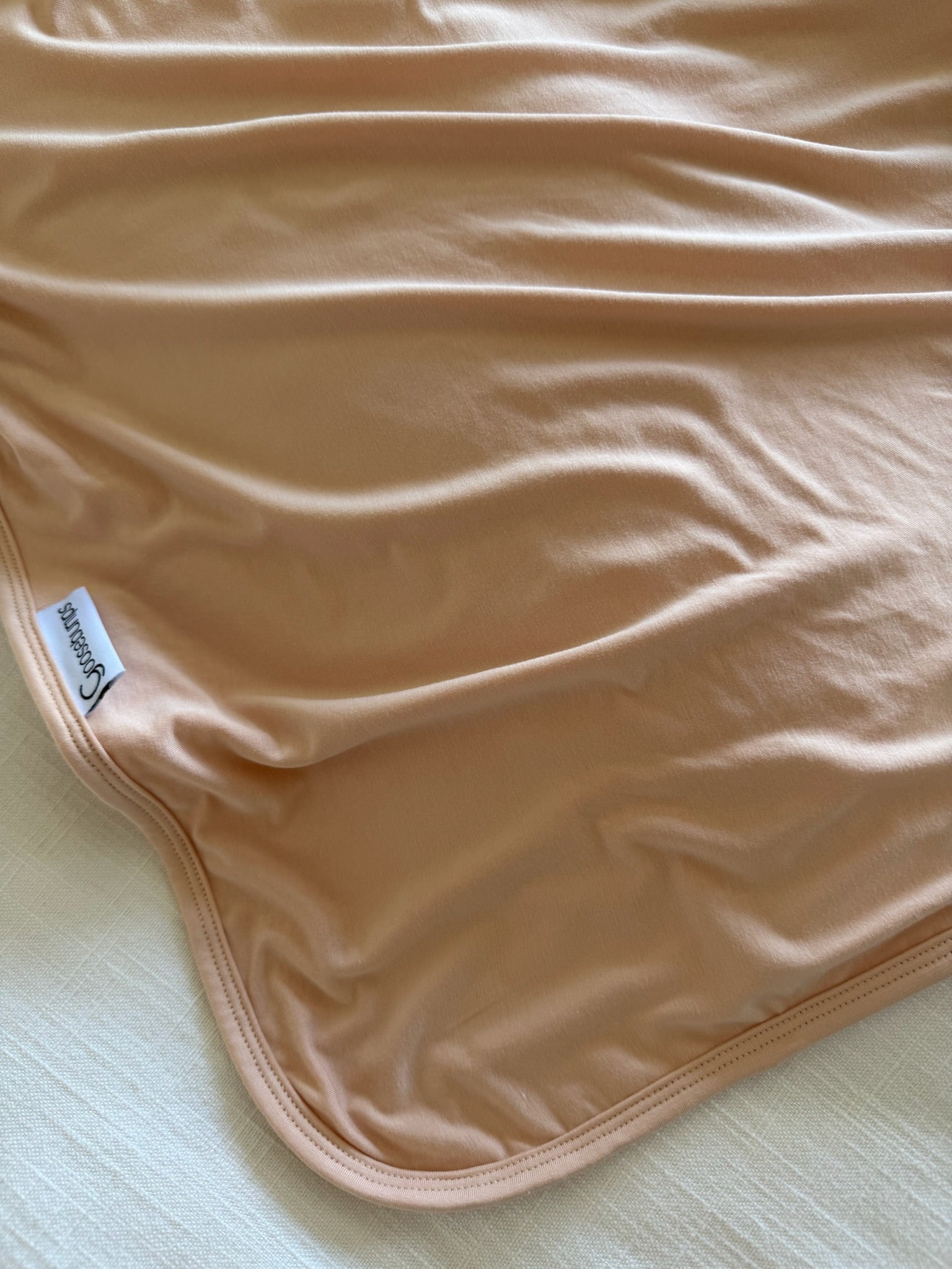 A close-up of peach-colored whisp bamboo loungewear fabric shows its smooth, slightly shiny texture with gentle wrinkles. A white tag with the brand name Cossac appears on one edge as the fabric rests on a light surface.