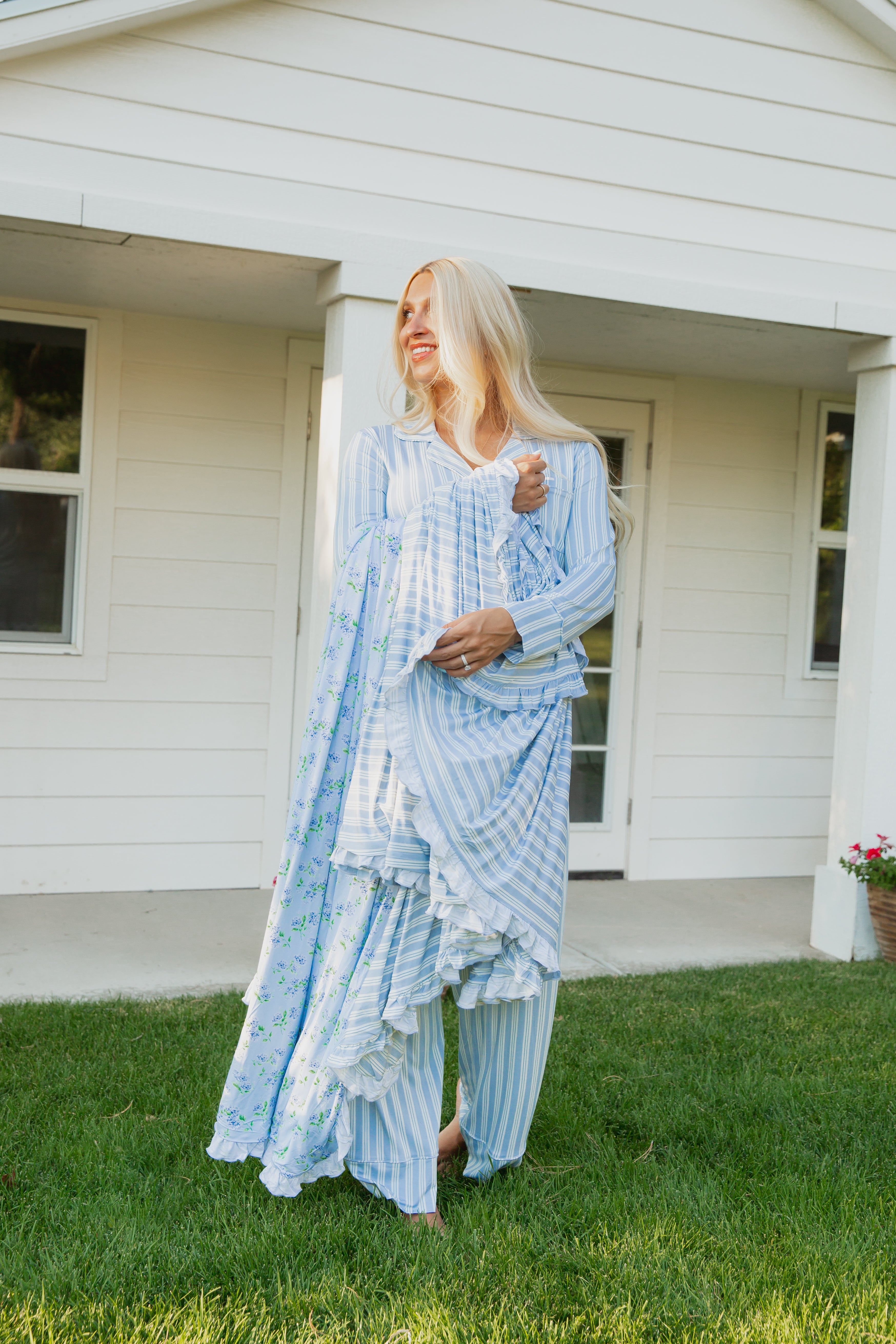 A woman with long blonde hair, barefoot on the grass in front of a white house, smiles and looks to the side as she gently wraps herself in the luxurious hydrangea / ruffle bamboo blanket, draped over her blue and white striped traditional outfit.